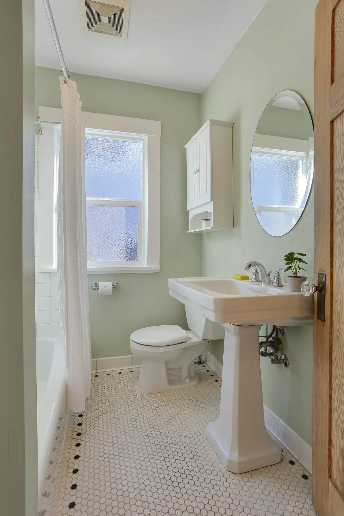 A small bathroom with a white toilet and pedestal sink, a white wall-mounted cabinet, a mirror, a window with frosted glass, a shower with a curtain, and a small potted plant on the sink. The floor has small hexagonal tiles.