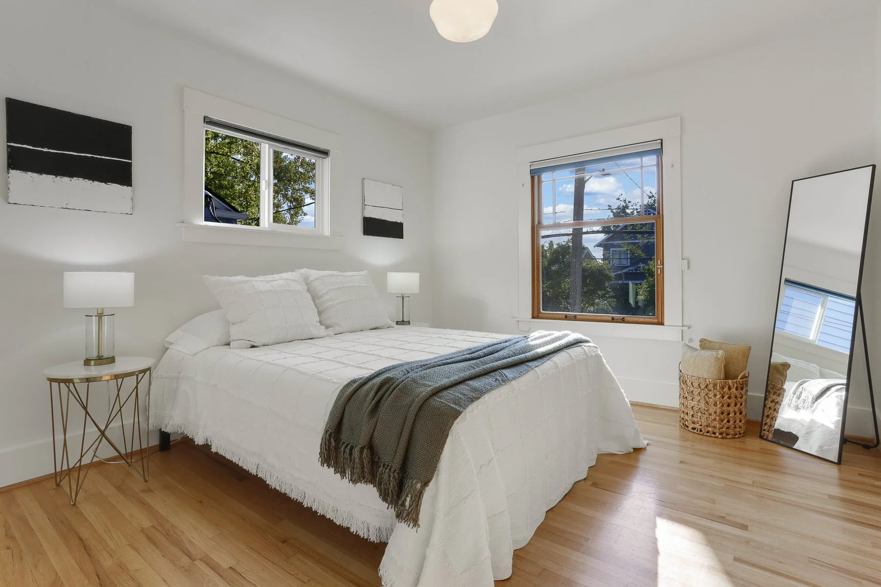 Bright bedroom with white walls, a bed with white bedding and pillows, a gray throw blanket, two white lamps on side tables, hardwood floors, two windows with trees outside, a standing mirror, and a wicker basket.