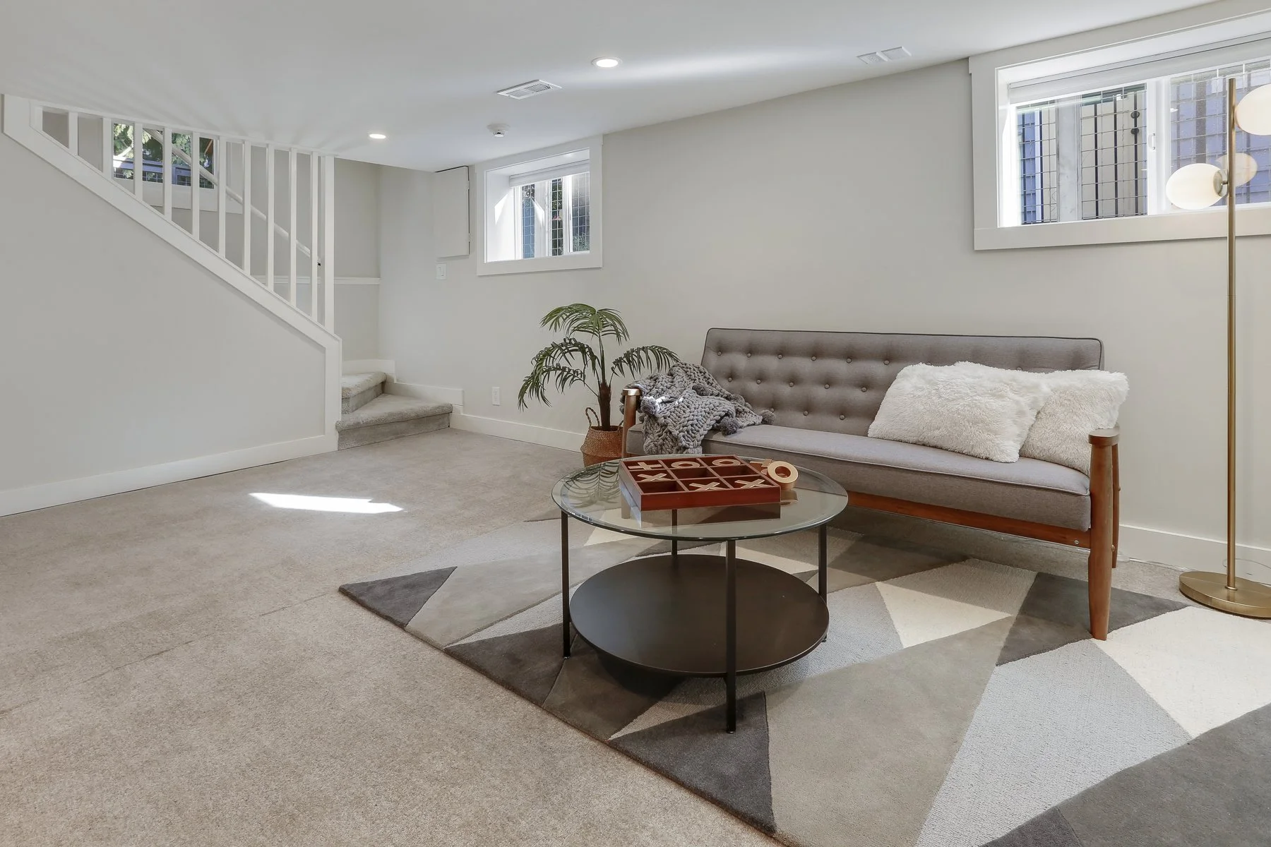 Living room with a gray tufted sofa, plush pillows, a glass coffee table with game pieces, a potted plant, and a floor lamp. The room has two small windows and a staircase with white railing.