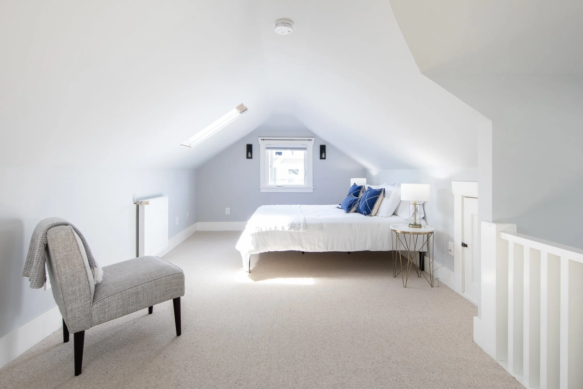 A bright and minimal bedroom with white walls and beige carpet. There's a bed with white bedding and blue pillows, a small round side table with a lamp, a gray armchair with a throw blanket, a window with white trim, and a ceiling-mounted smoke detec