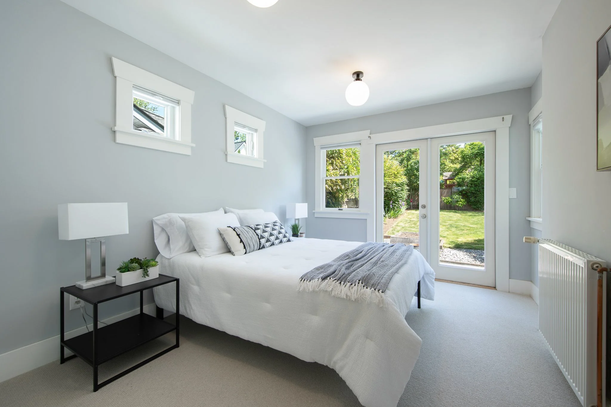 A bright bedroom with a large window and glass door overlooking a garden. The bed has white bedding and patterned pillows, with a nightstand and lamps on either side. White trim decorates the light gray walls.