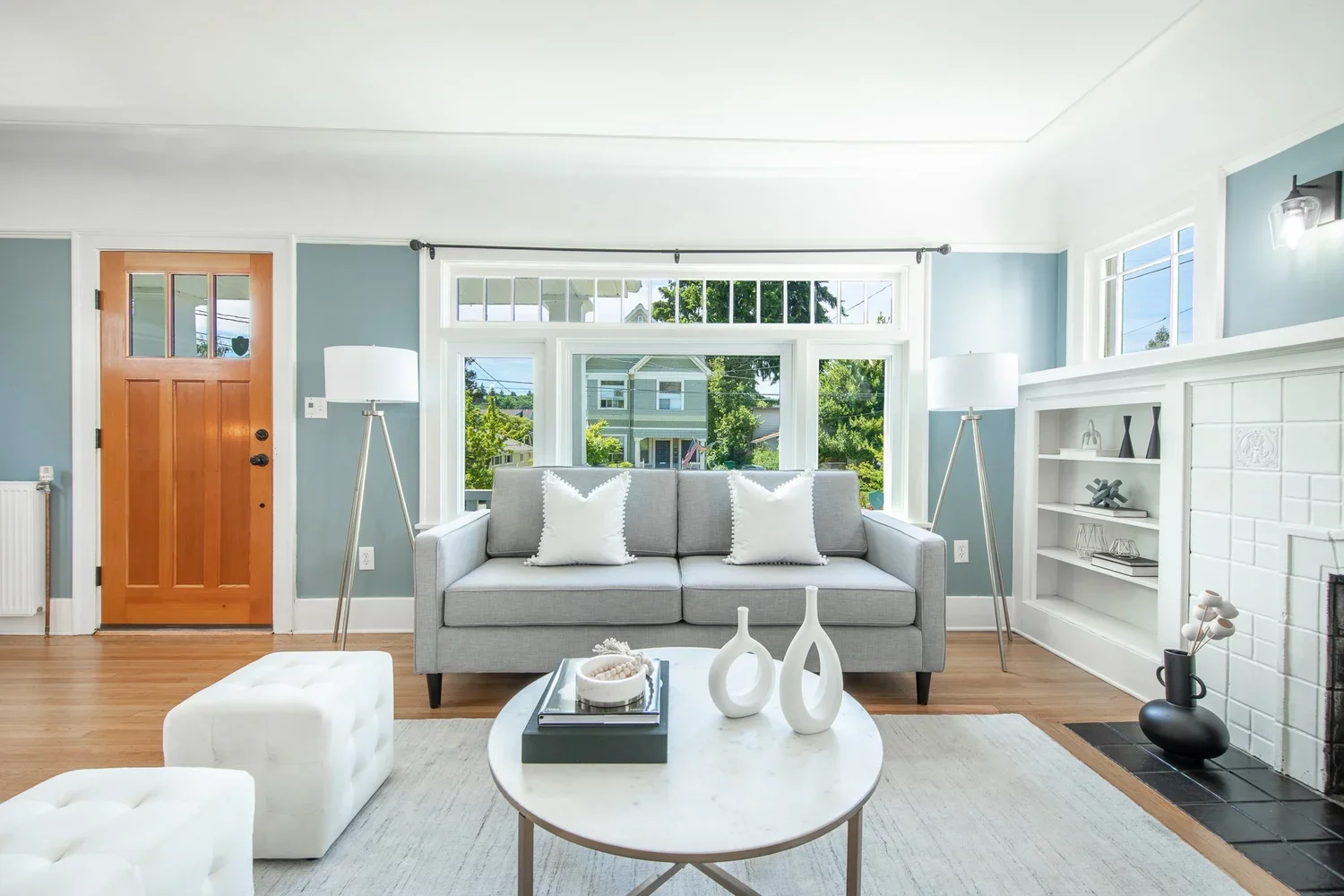 Bright living room with a gray sofa, white pillows, and large front windows showing a neighborhood scene; includes two tall floor lamps and decorative vases on a coffee table.