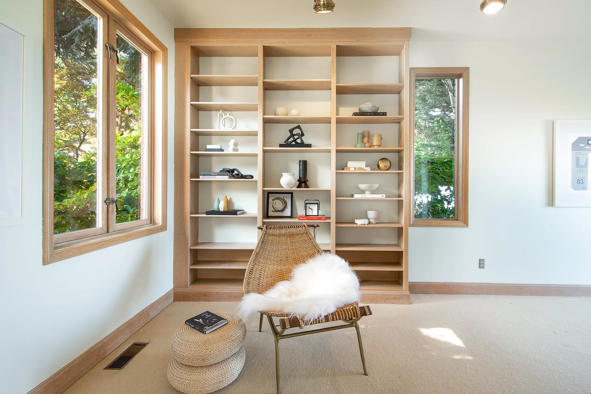 Room with light-colored walls, large windows showing greenery outside, and a tall wooden shelving unit filled with decorative objects. There is a woven chair with a fur throw, a small round wicker ottoman with a book on top, and a beige carpeted floo