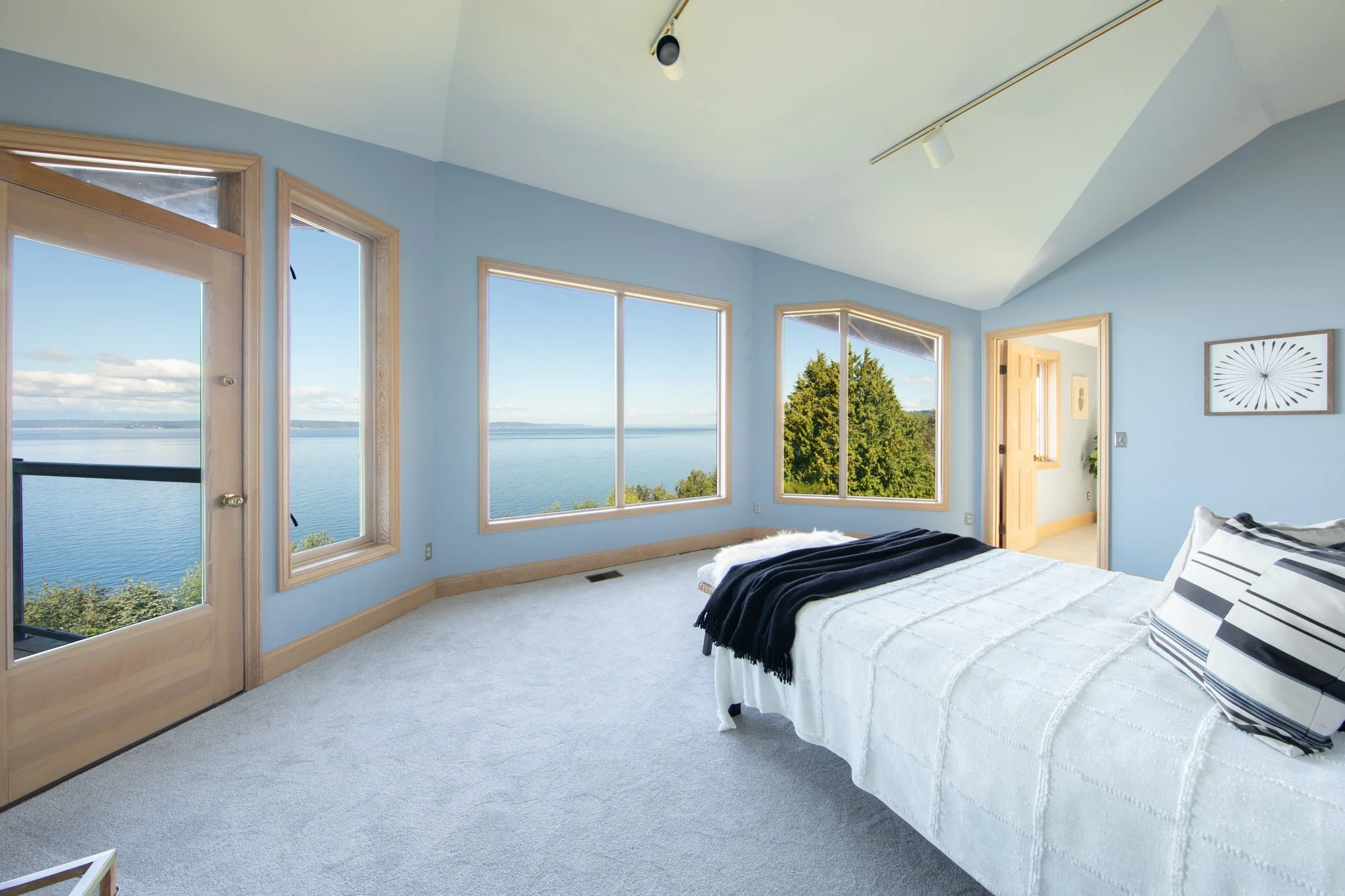 A bedroom with light blue walls and large windows overlooking a lake and trees. There is a bed with white bedding and black and white striped pillows. The room has wooden window frames, a wooden door, artwork on the wall, and a ceiling light fixture.