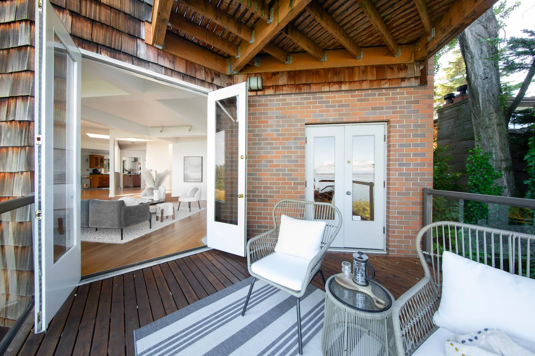 Balcony with outdoor seating, including white chairs and a small table, overlooking a living room with brick and wood interior walls.