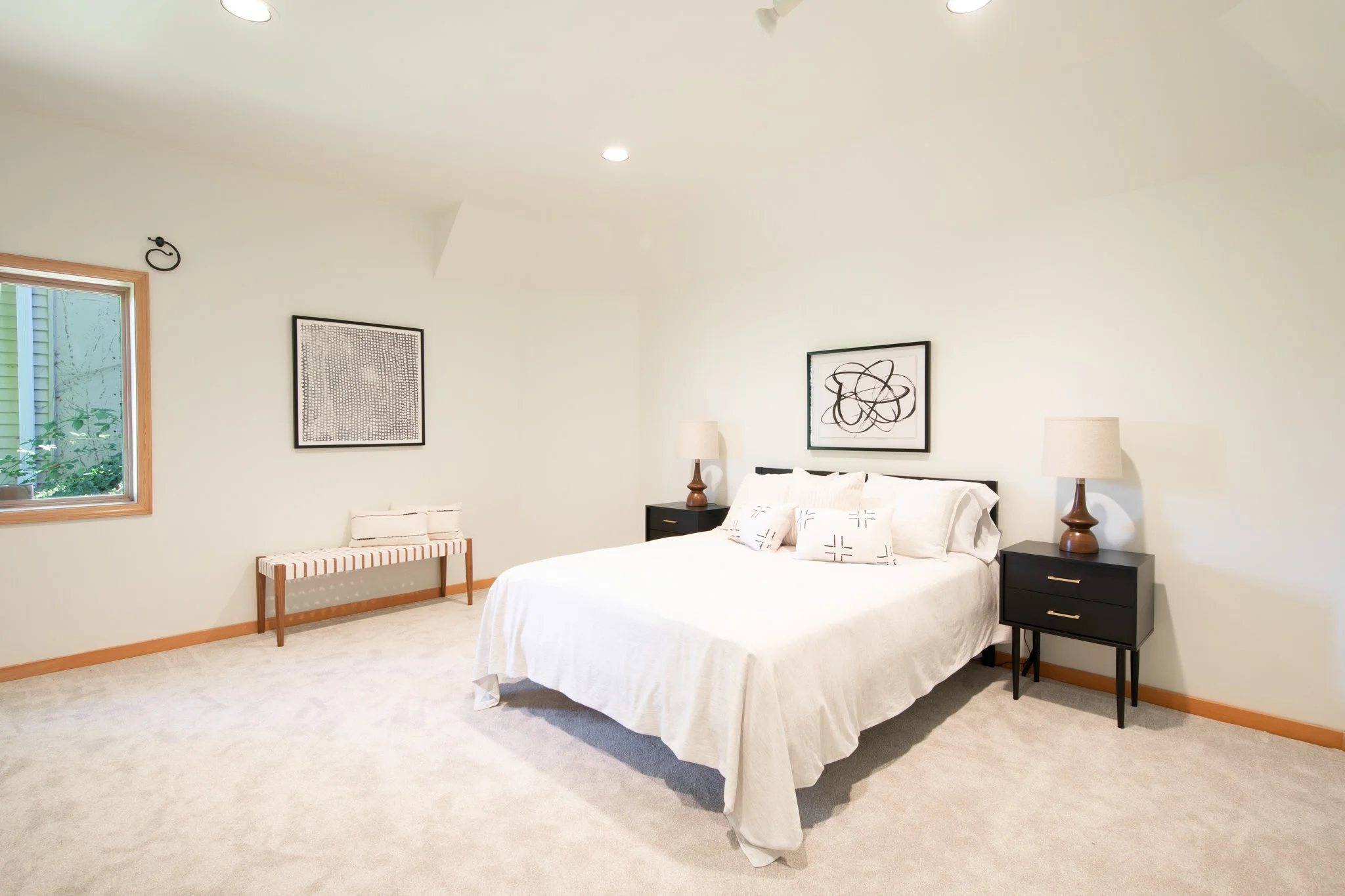 A bedroom with a white bed, black nightstands, and matching lamps. Decor includes two modern black and white wall art pieces. There is a window with a wooden frame and beige carpeted flooring.