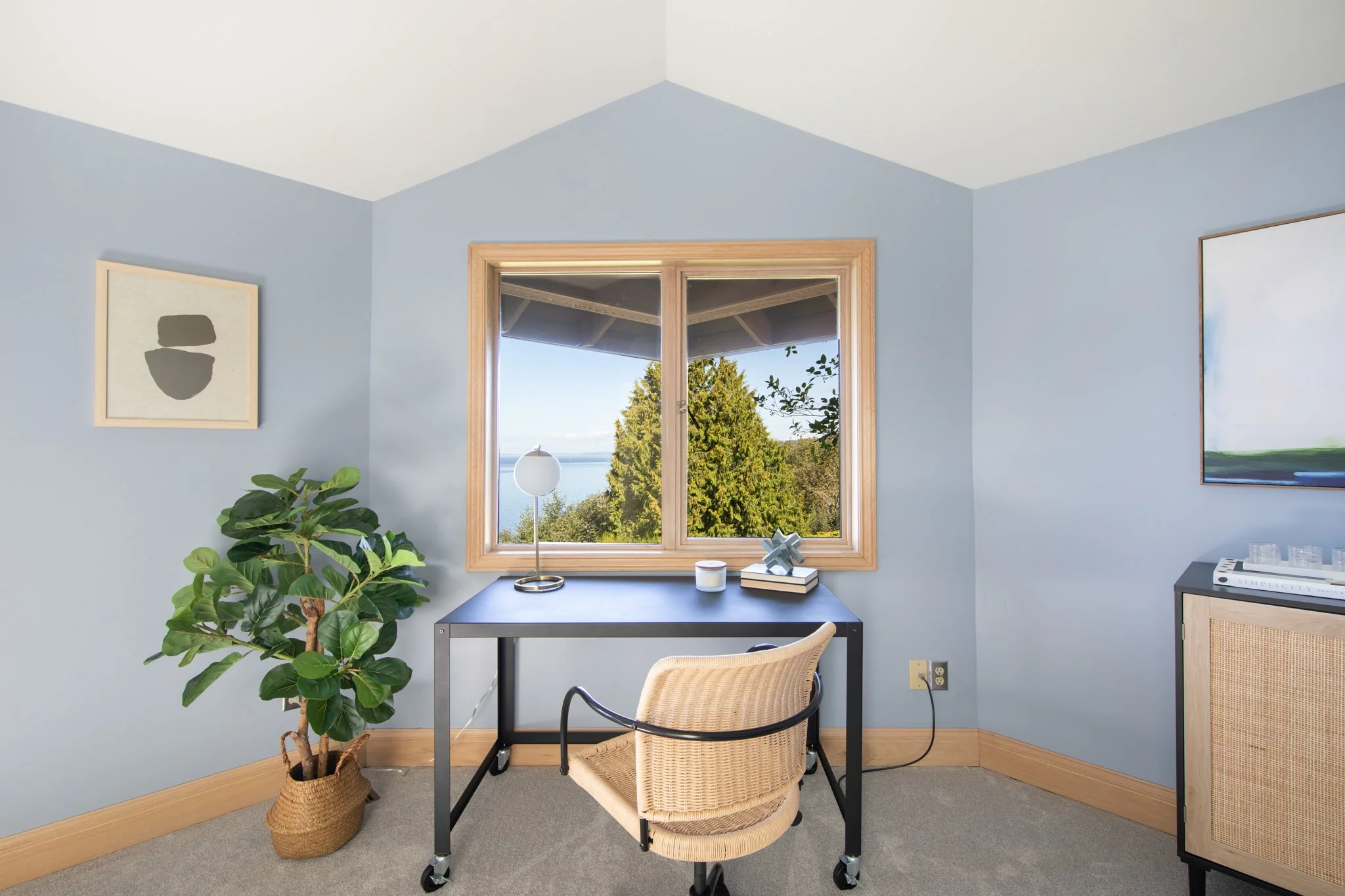A home office with light blue walls, a window showing trees and water outside, a desk with a lamp, a cartoon-like figurine, and books, a woven chair, artwork on the walls, and a potted plant on the floor.