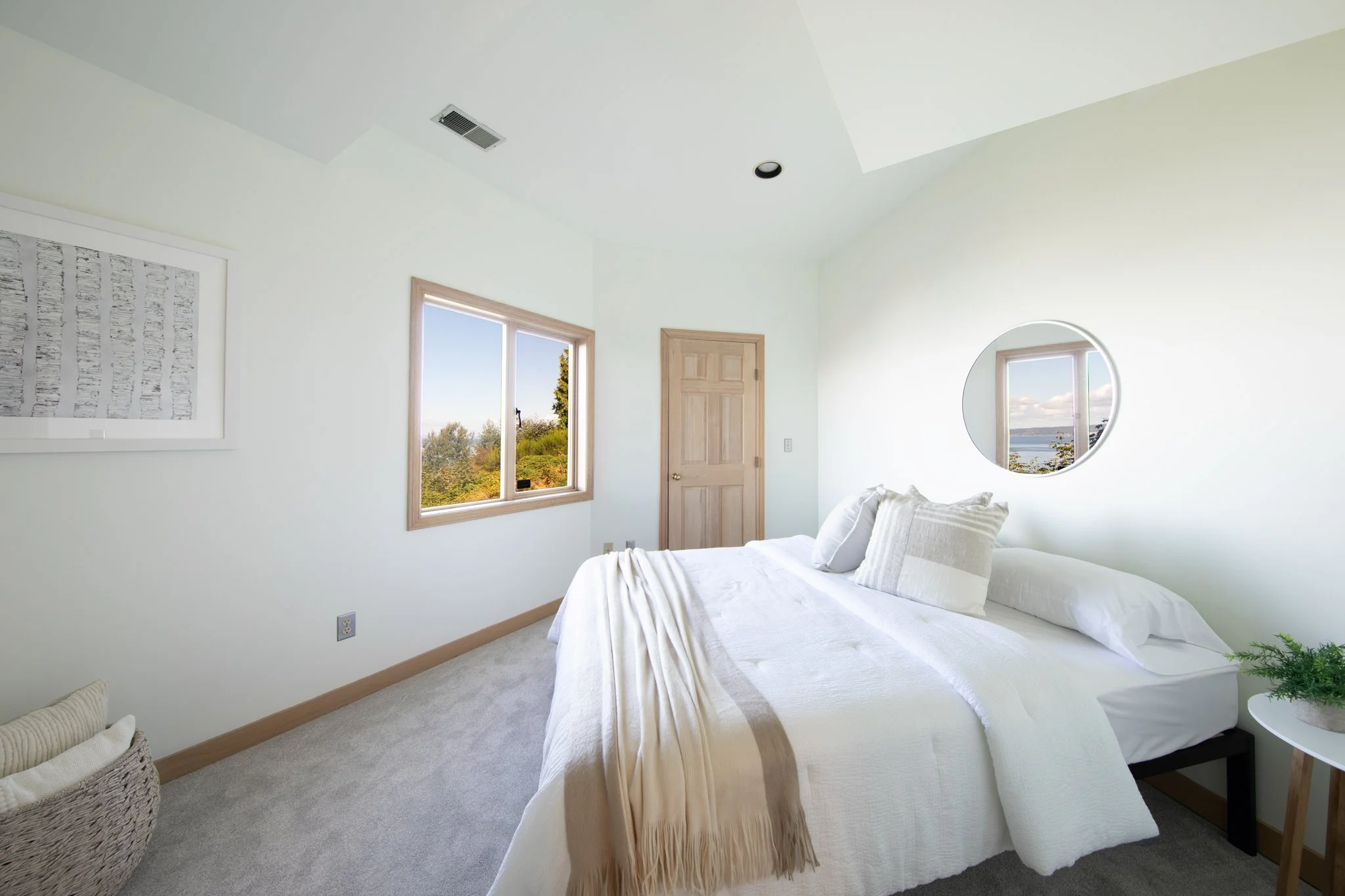 A bright bedroom with white walls, a bed with white linens and pillows, two windows showing a scenic outdoor view, a wooden door, a framed black-and-white artwork, a round mirror reflecting the window view, and a potted plant.
