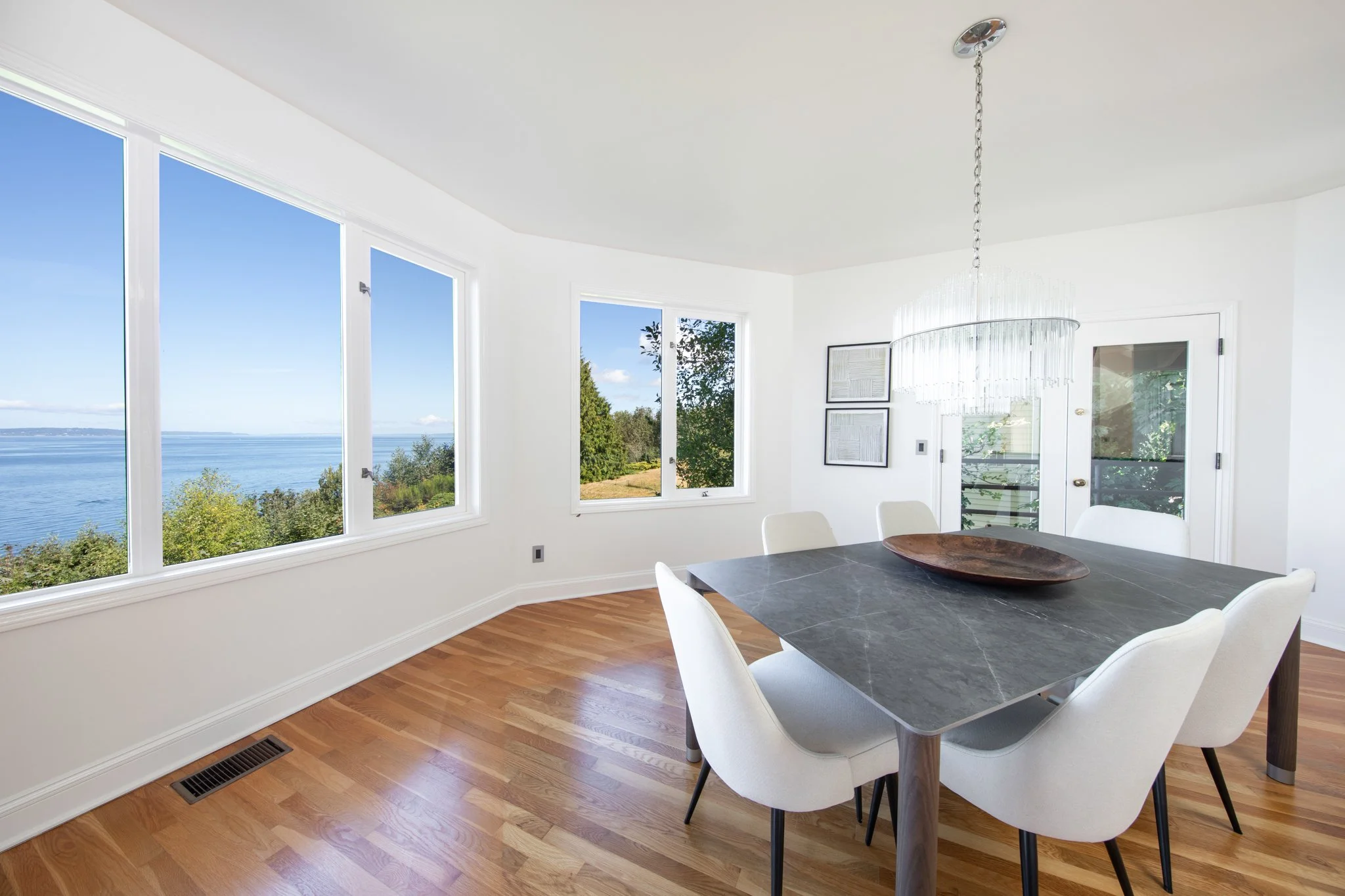 Bright dining room with large windows overlooking a body of water and greenery, white walls, hardwood floor, a dark gray table, and white chairs.