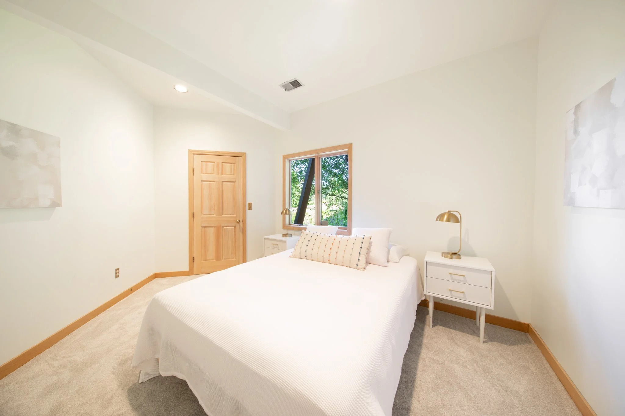 A bedroom with white walls, a bed with white bedding and pillows, two white nightstands with gold lamps, a window showing greenery outside, and a wooden door.