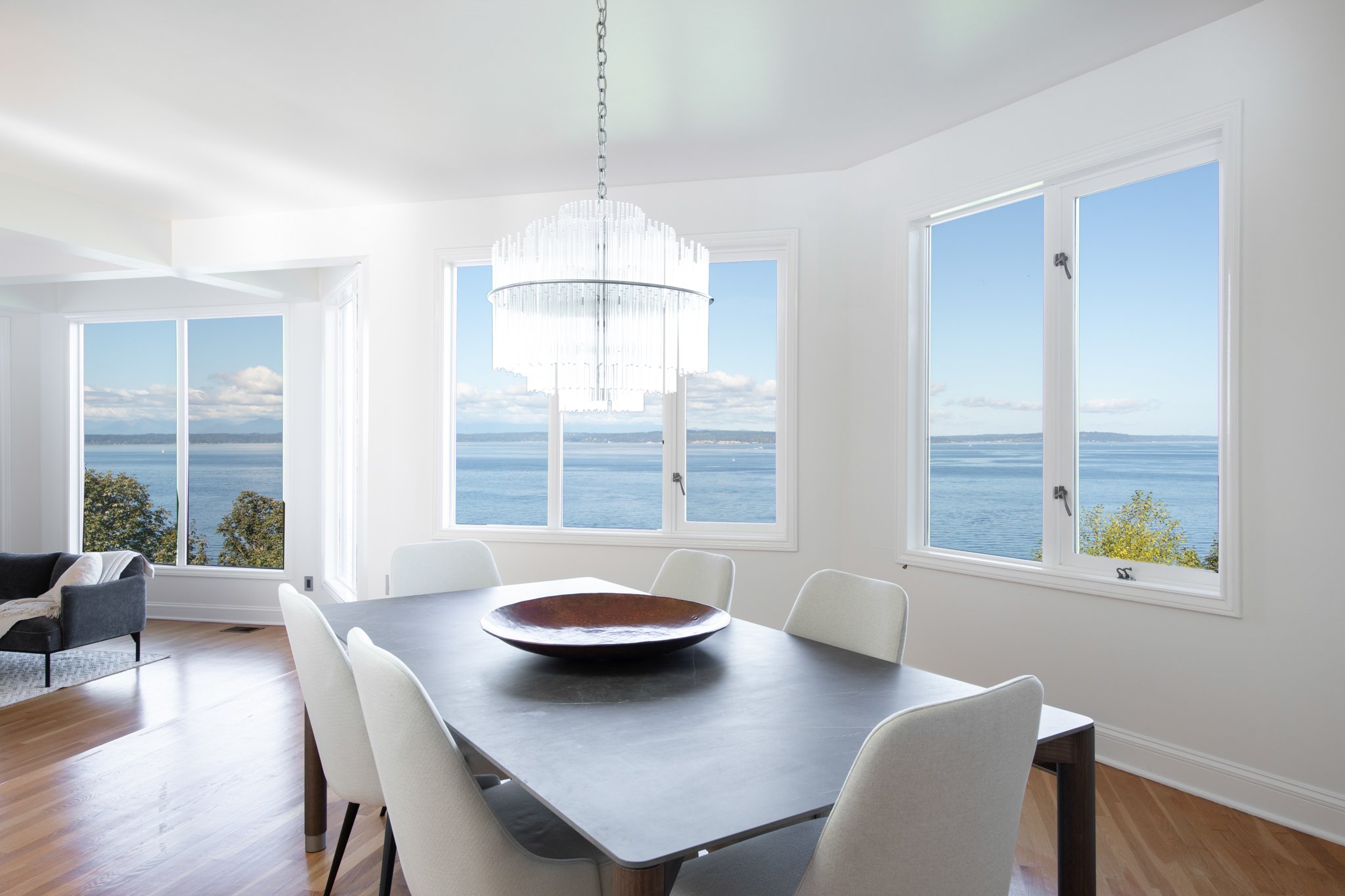 Interior view of a modern dining area with large windows showing a body of water and sky outside, a chandelier hanging from the ceiling, a round dining table with a decorative bowl, and chairs surrounding the table.