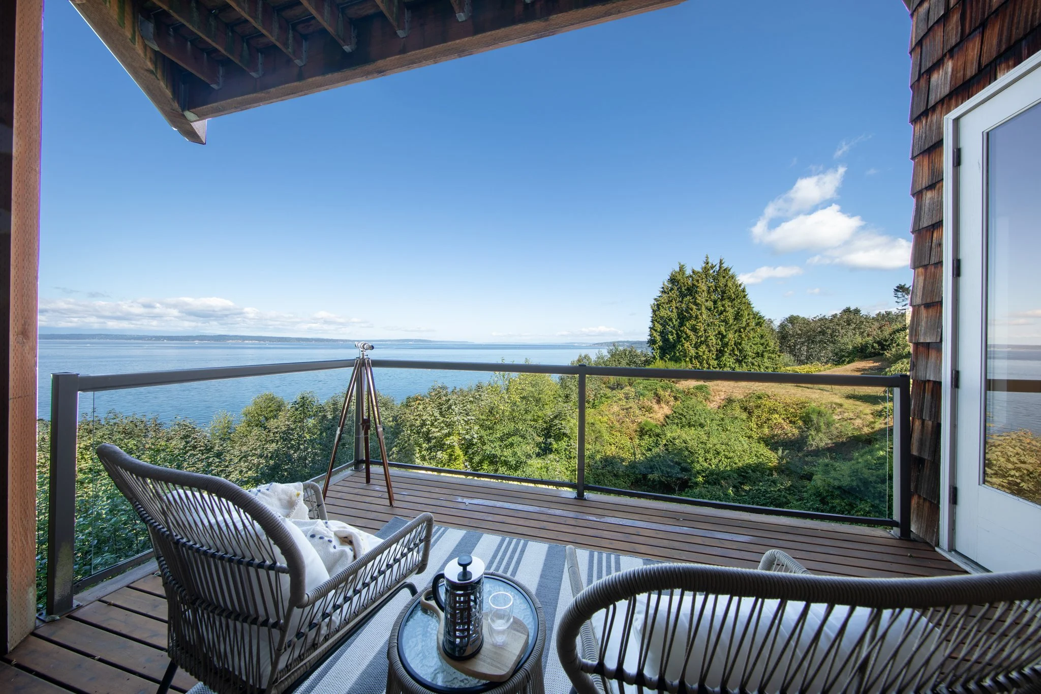 Balcony overlooking a body of water with trees and a blue sky with clouds, outdoor furniture including a small round table, chairs, and a telescope