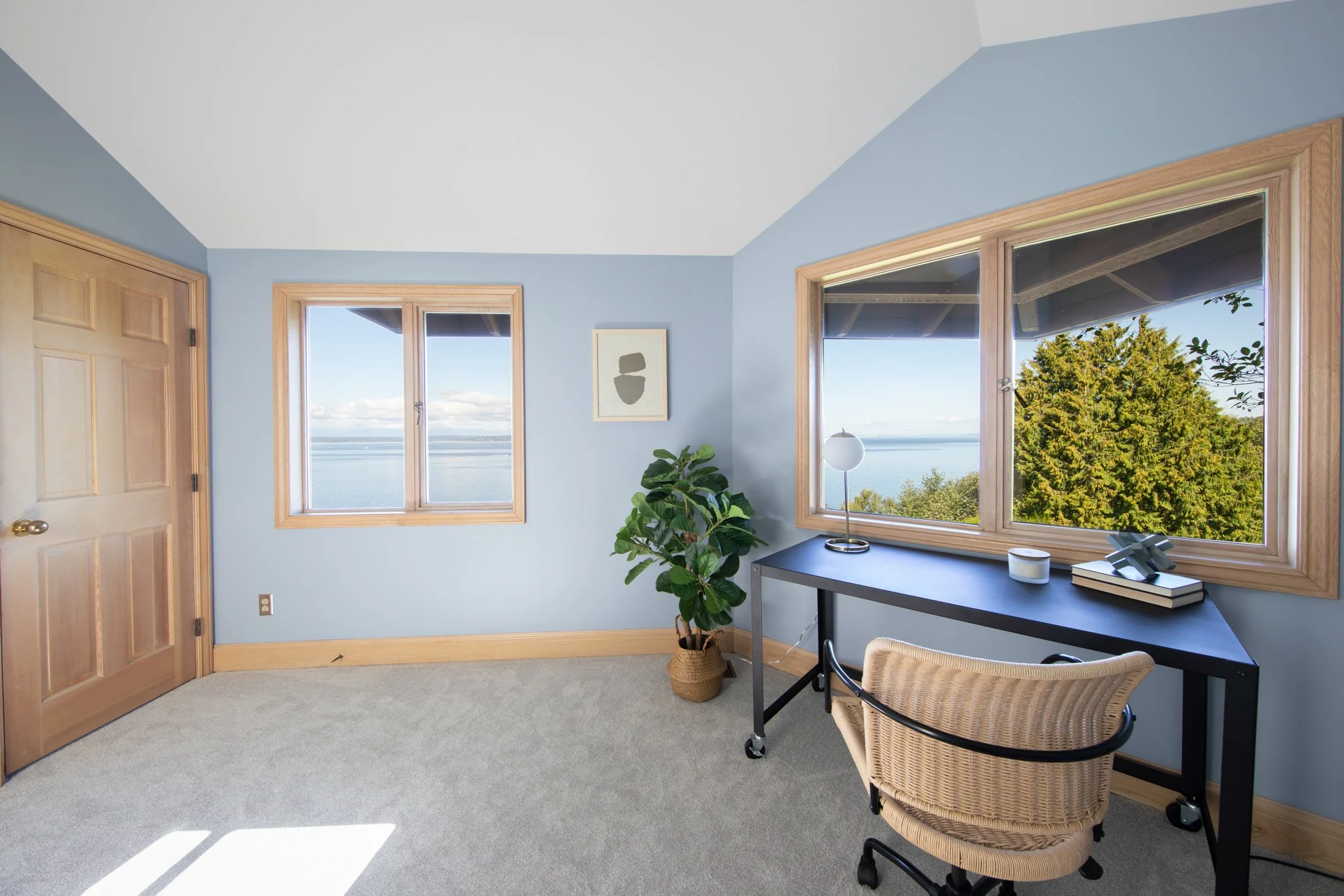 A cozy room with light blue walls and large windows showing a scenic view of water and trees, decorated with a framed abstract artwork, a potted plant, a modern table lamp, and a desk with books and decorative items.