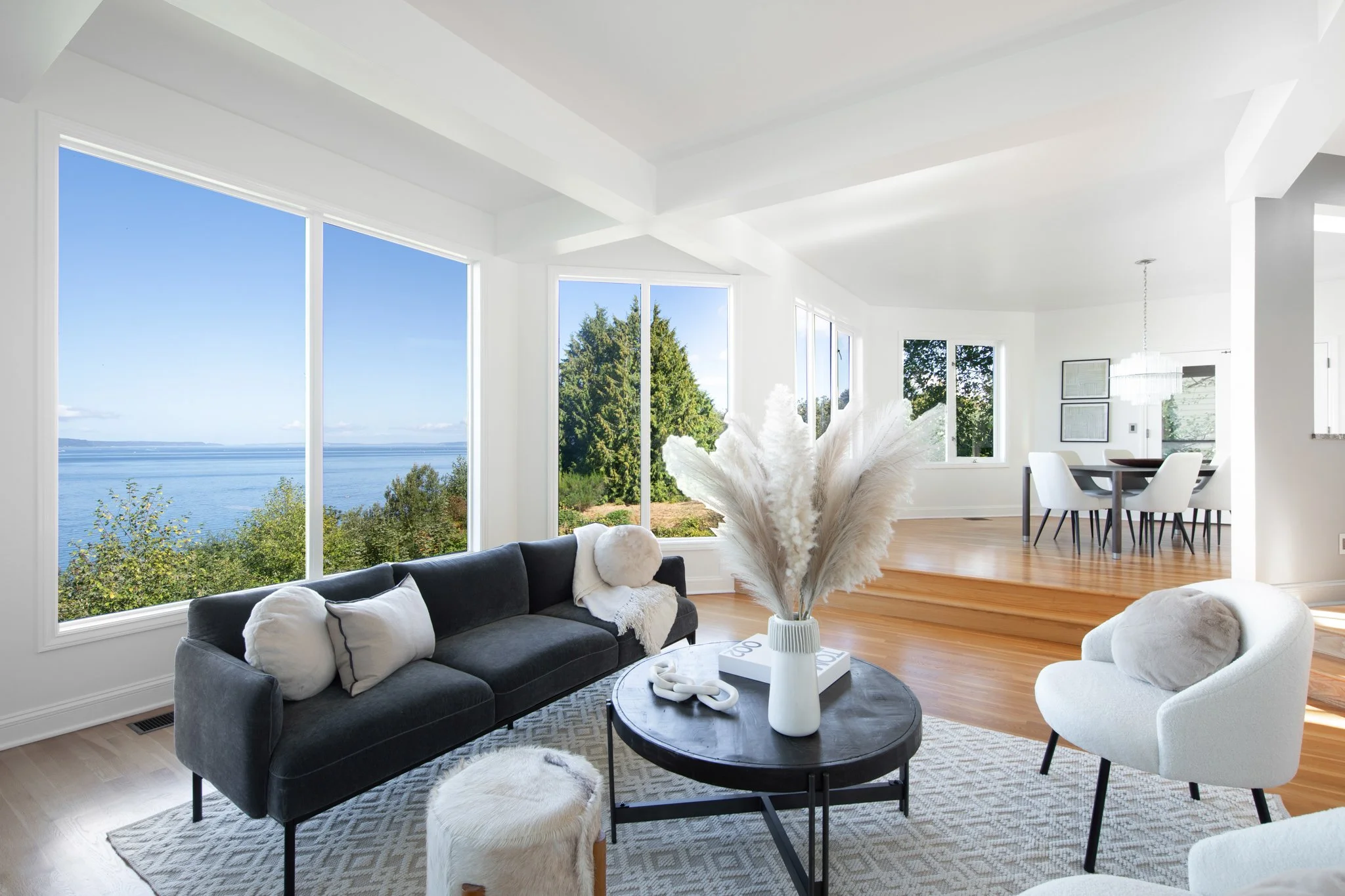 A modern living room with large windows overlooking a body of water and trees, decorated with a black sofa, white and beige pillows, round ottomans, a round coffee table with white vases, and a floral arrangement, with a dining area visible in the ba