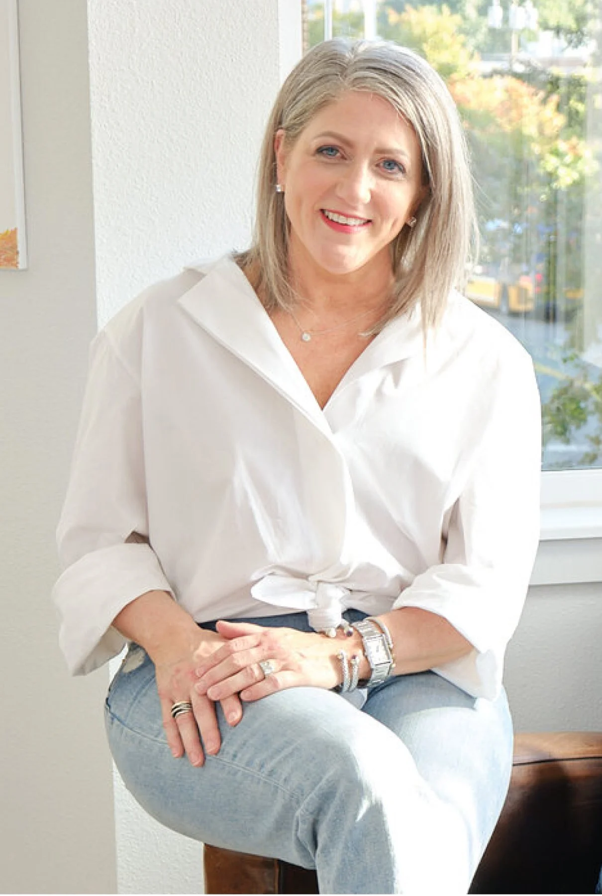 A woman with shoulder-length gray hair, wearing a white blouse, light blue jeans, and jewelry, sitting on a wooden stool by a window with a view of trees and a street outside, smiling at the camera.