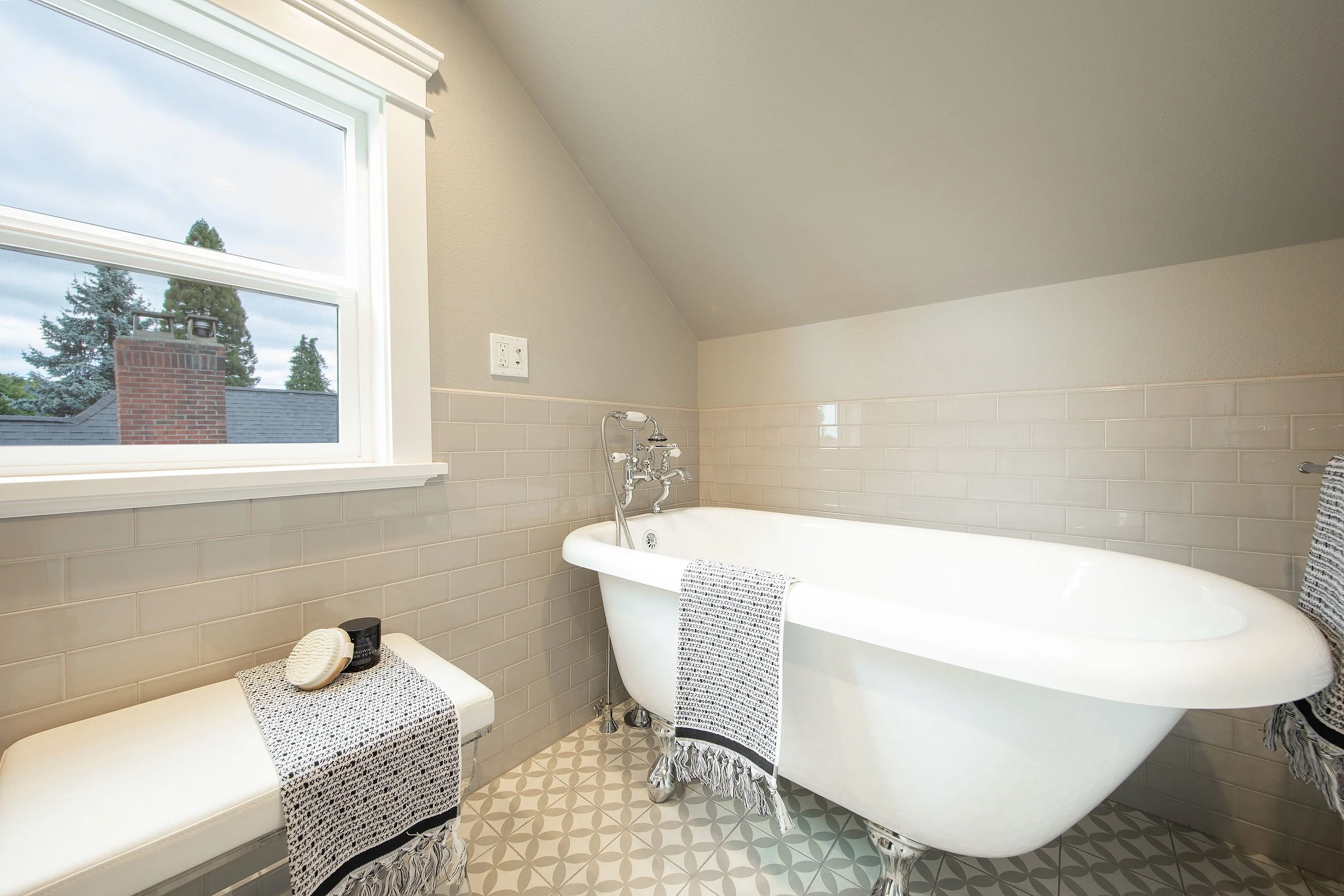 A bathroom with a clawfoot bathtub next to a window showing a view of trees and a brick chimney. The bathtub has striped towels hanging over the sides, and there is a towel and some toiletries on a small bench nearby.