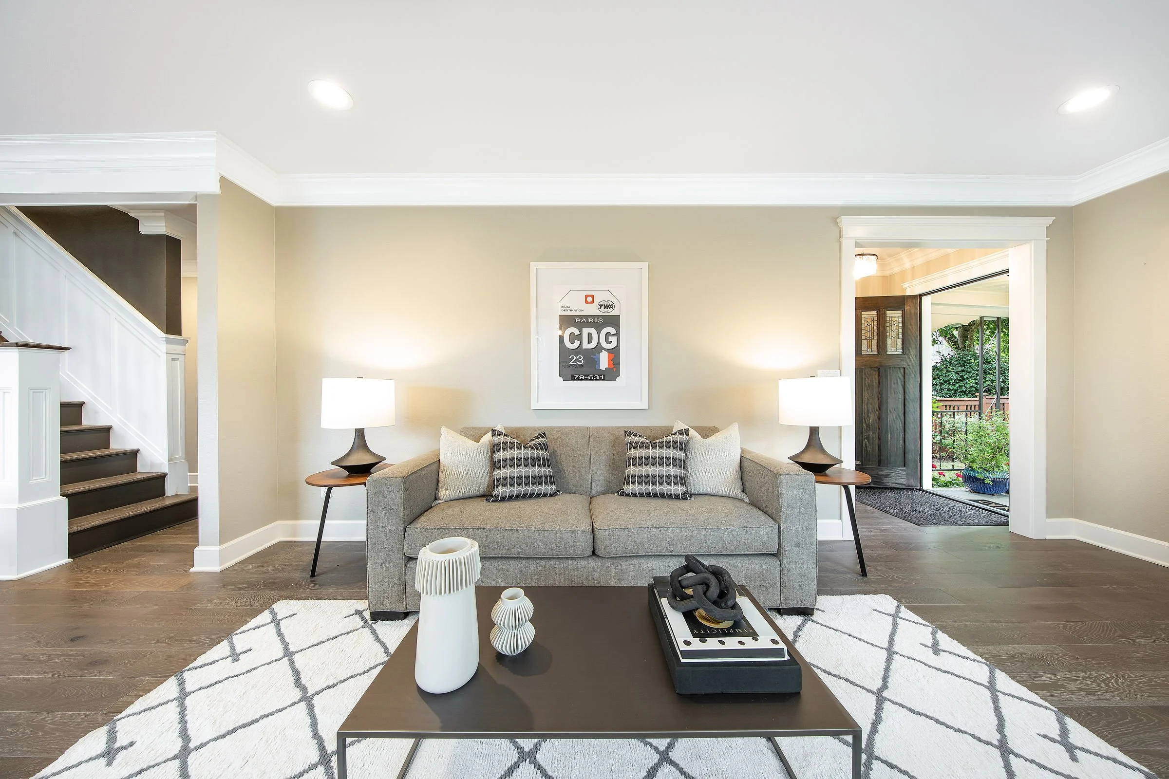 Living room with beige walls, white crown molding, and a beige sofa with patterned pillows. There are two table lamps on either side of the sofa, a coffee table with decorative items, and a framed art piece on the wall. To the left, a staircase with 