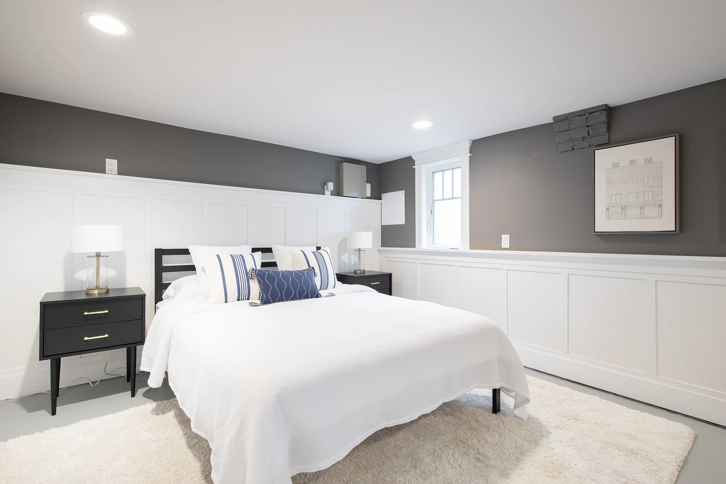 A bedroom with a white bed, black nightstands, white lamps, gray walls, framed artwork, a window, and a white carpet.