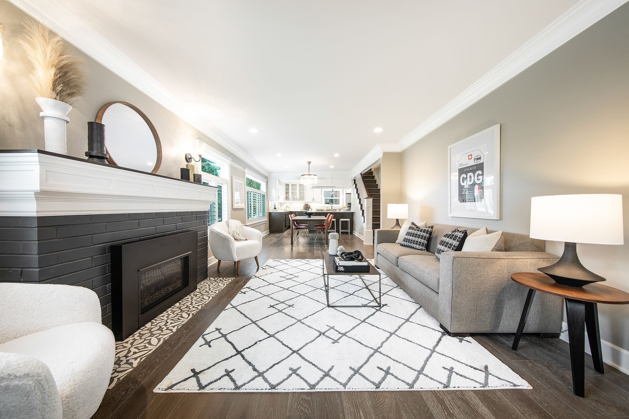 A spacious, modern living room with a black fireplace, white walls, and dark wood flooring. The room features a large light gray sofa with patterned cushions, a white armchair, and a glass coffee table with decorative items. Artworks and a framed pos