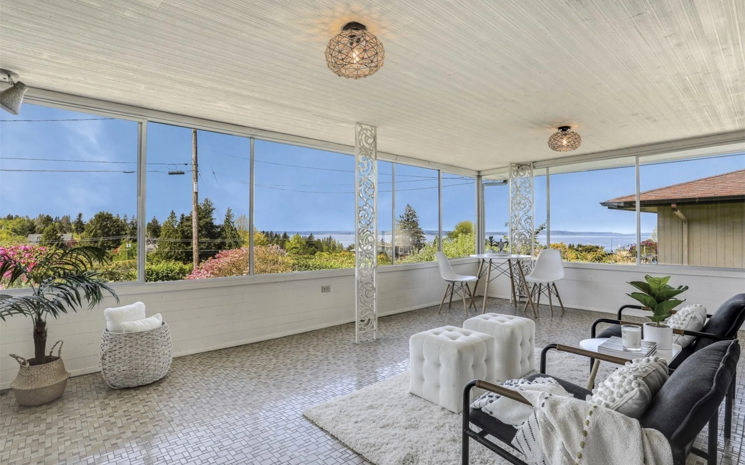 Bright sunroom with large windows showing a view of trees, water, and a partly cloudy sky. The room has hanging wicker light fixtures, white and black furniture, plants, and decorative interior accents.
