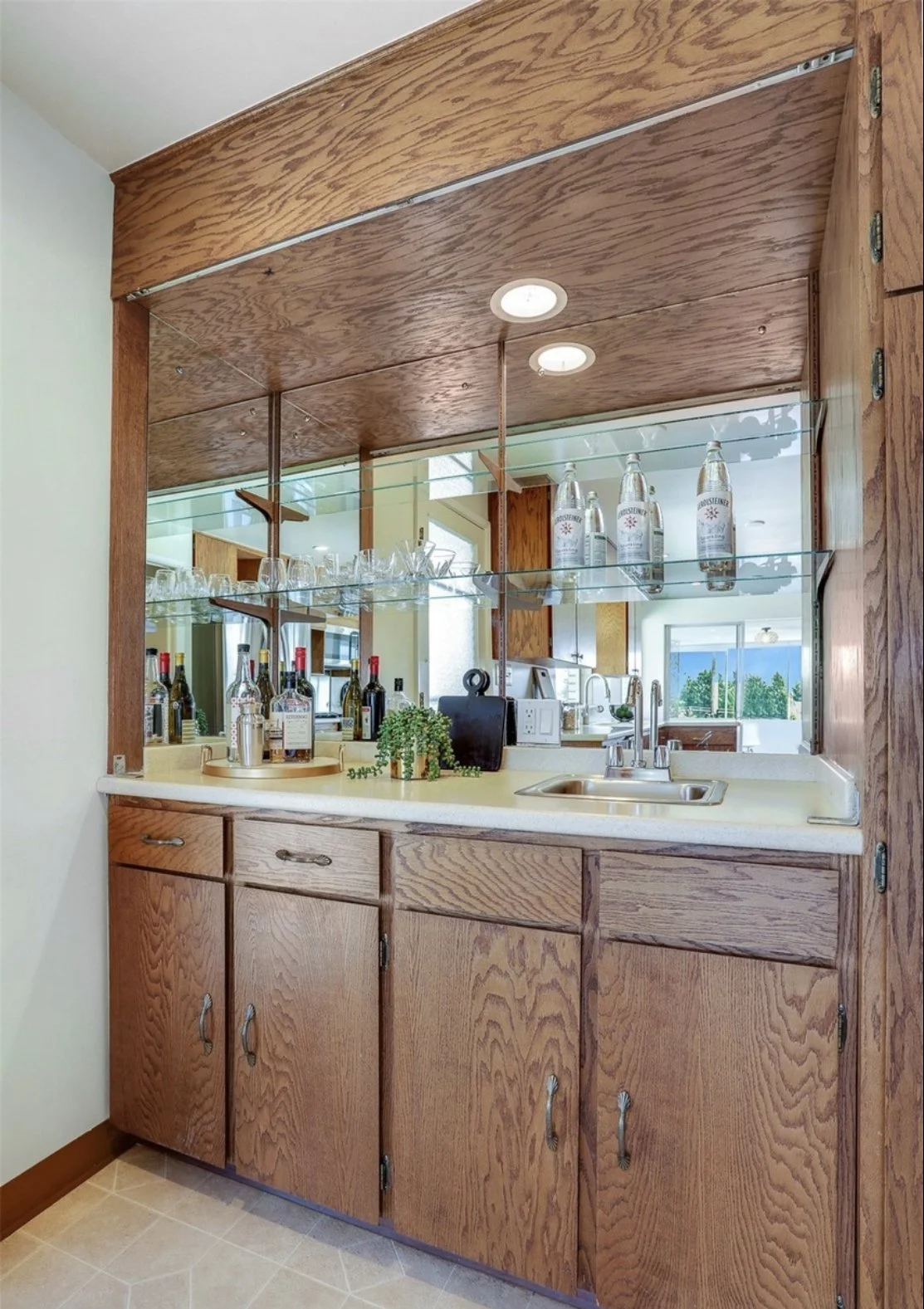 A wooden home bar with a large mirror, glass shelves, various liquor bottles, glasses, and a small sink.