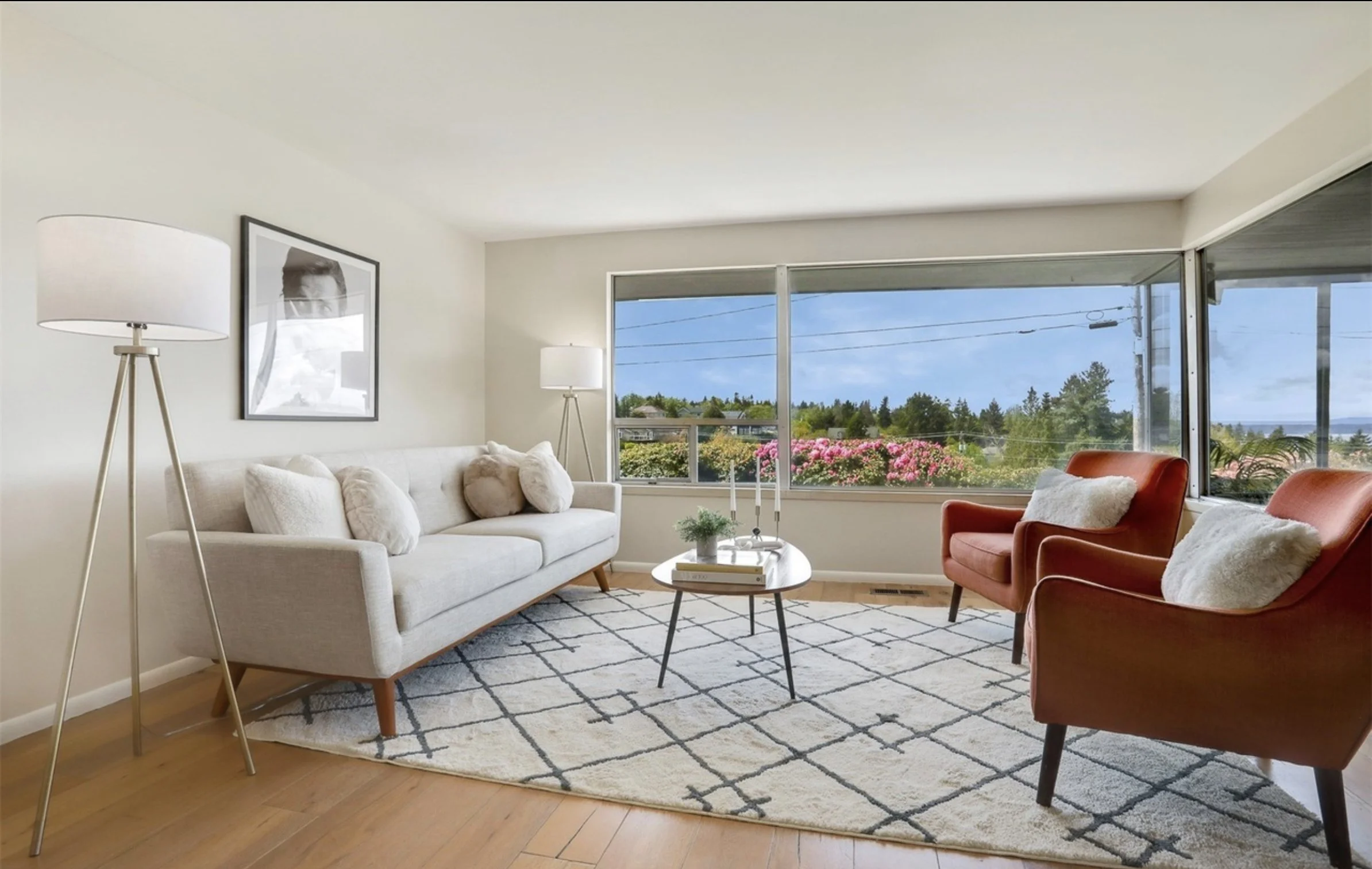 A living room with a white sofa, two orange chairs, and a black and white framed portrait on the wall. Large windows show a view of trees, flowers, and power lines outside. There are white pillows on the sofa and chairs, a round coffee table with dec