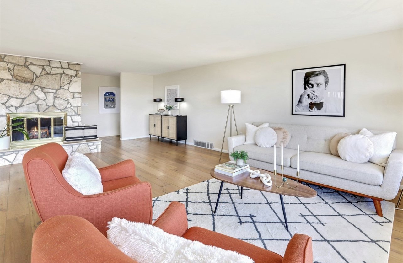 Living room with a stone fireplace, white sofa with round pillows, orange armchairs with fluffy white pillows, a wooden coffee table with decor, and a black and beige sideboard against a white wall, with black and white framed photo hanging above the
