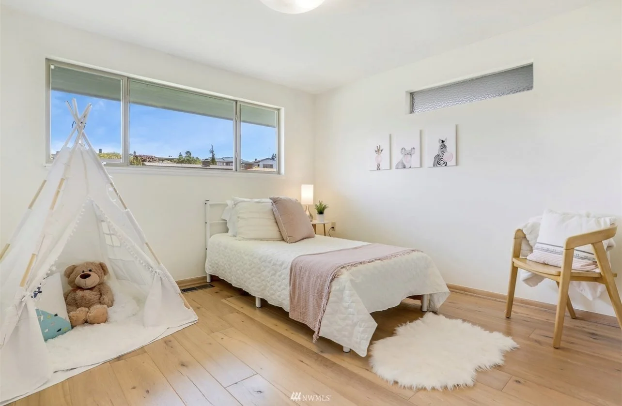 A bright, minimalist bedroom with a white bed, wooden floors, a small tent with teddy bear, a chair with blankets, and framed art of animals on the wall.