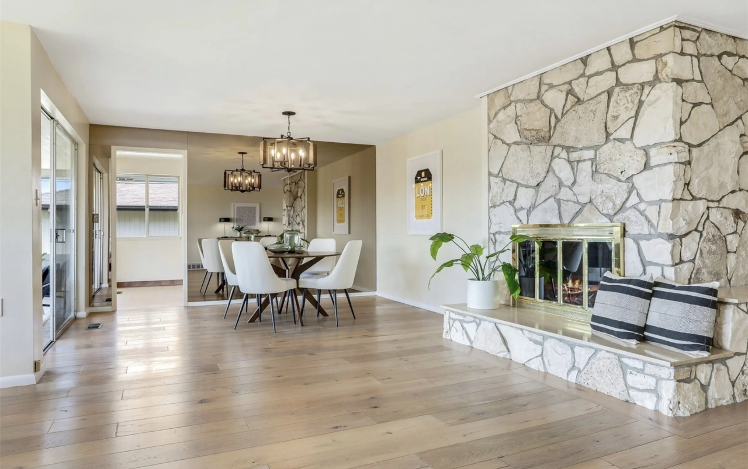 A living and dining room with a stone fireplace, wooden floors, and modern decor, featuring a potted plant near the fireplace and a dining table with white chairs.