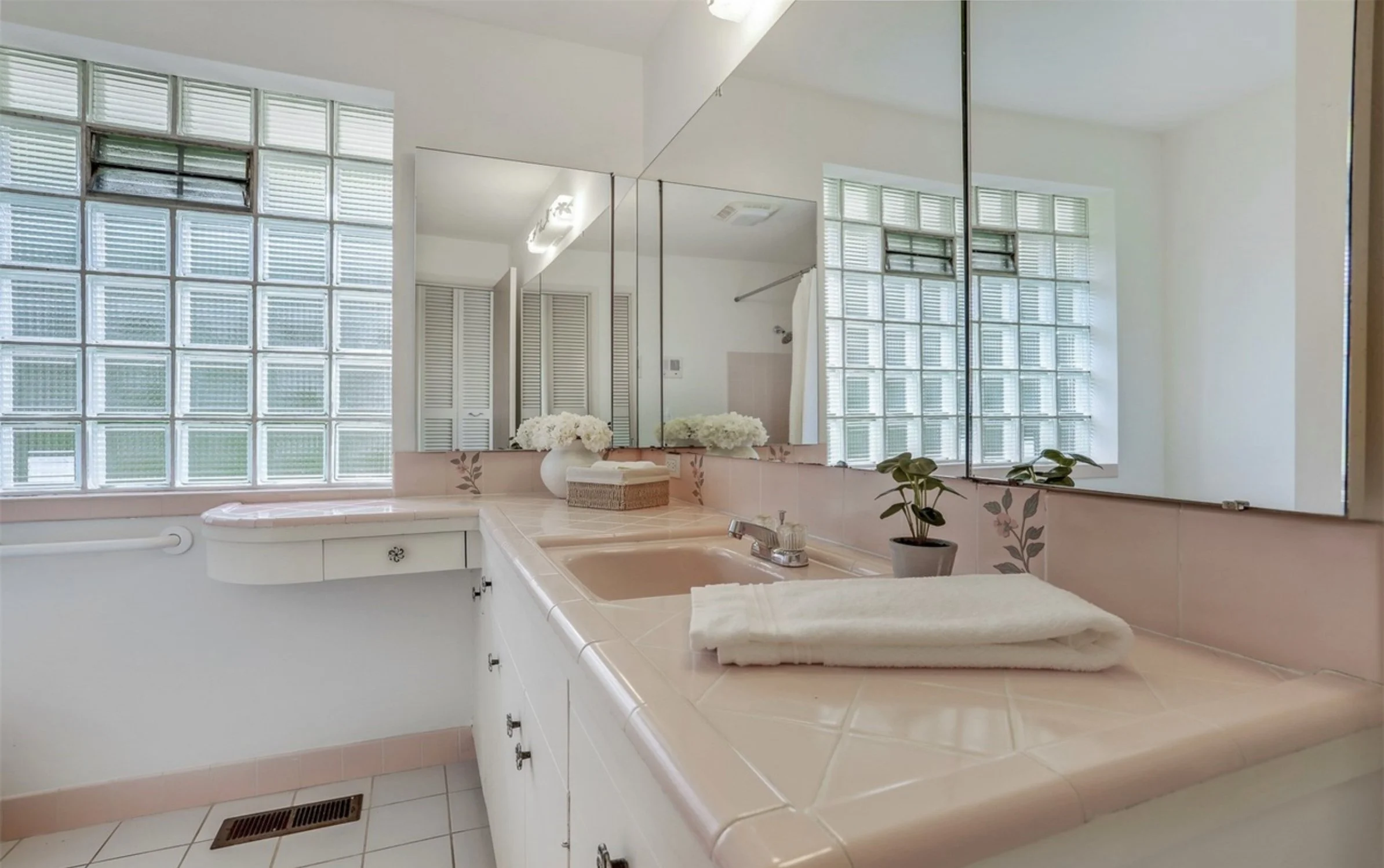 Bathroom with a large mirror, pink sink, white tiled countertop, and a large glass block window that lets in natural light.
