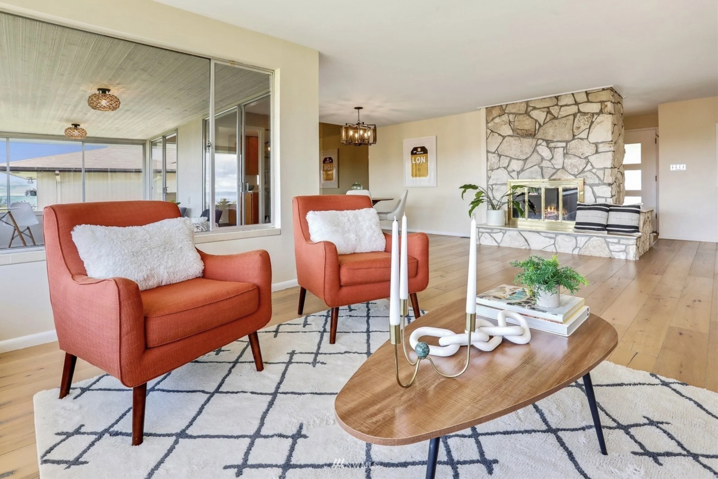 Living room with coral sofa chairs, a wooden coffee table, a patterned rug, and a fireplace with large stone surround, with a ceiling light fixture and sliding glass doors leading to an outdoor patio.