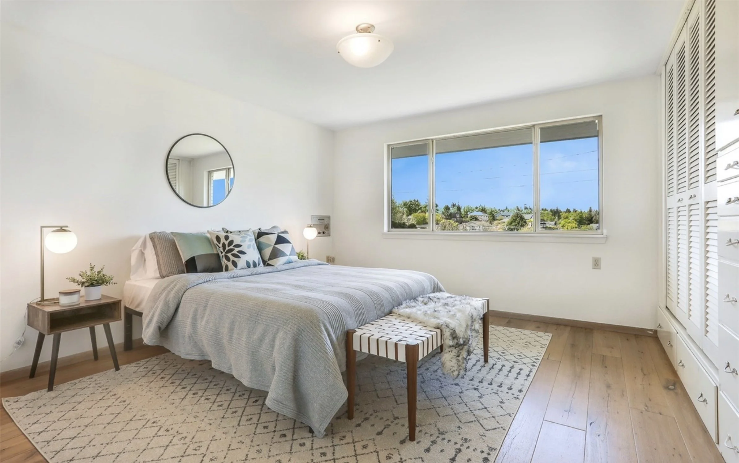 A bright bedroom with a large window showing a view of trees and houses outside, a bed with a patterned blanket and multiple pillows, bedside tables with lamps, a round mirror on the wall, and a closet with louvered doors.