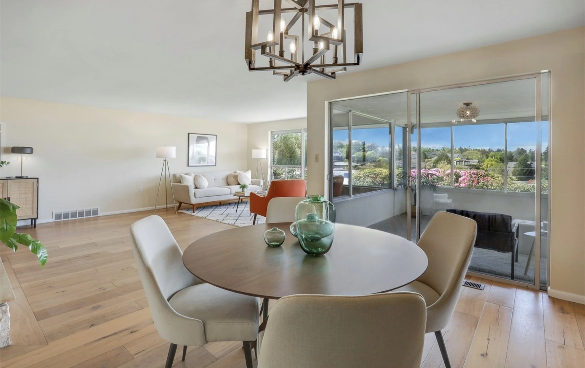 Interior of a modern living room and dining area with large windows and a view of trees and flowers outside, a round dining table with beige chairs, a white sofa, and various lamps.