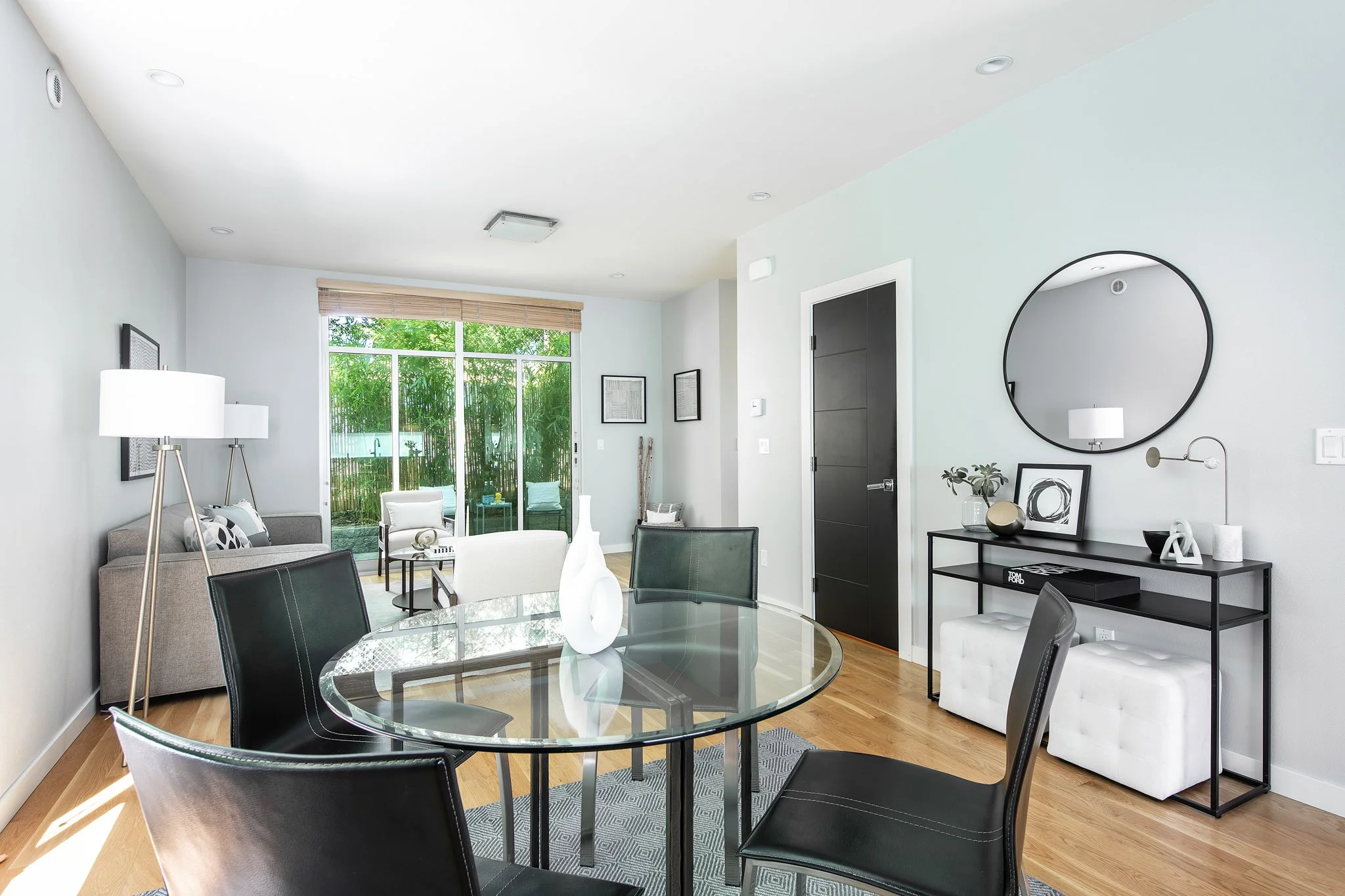 Modern living room with white walls, hardwood floors, black and white furniture, glass dining table, round mirror, and large window with bamboo blinds showing greenery outside.
