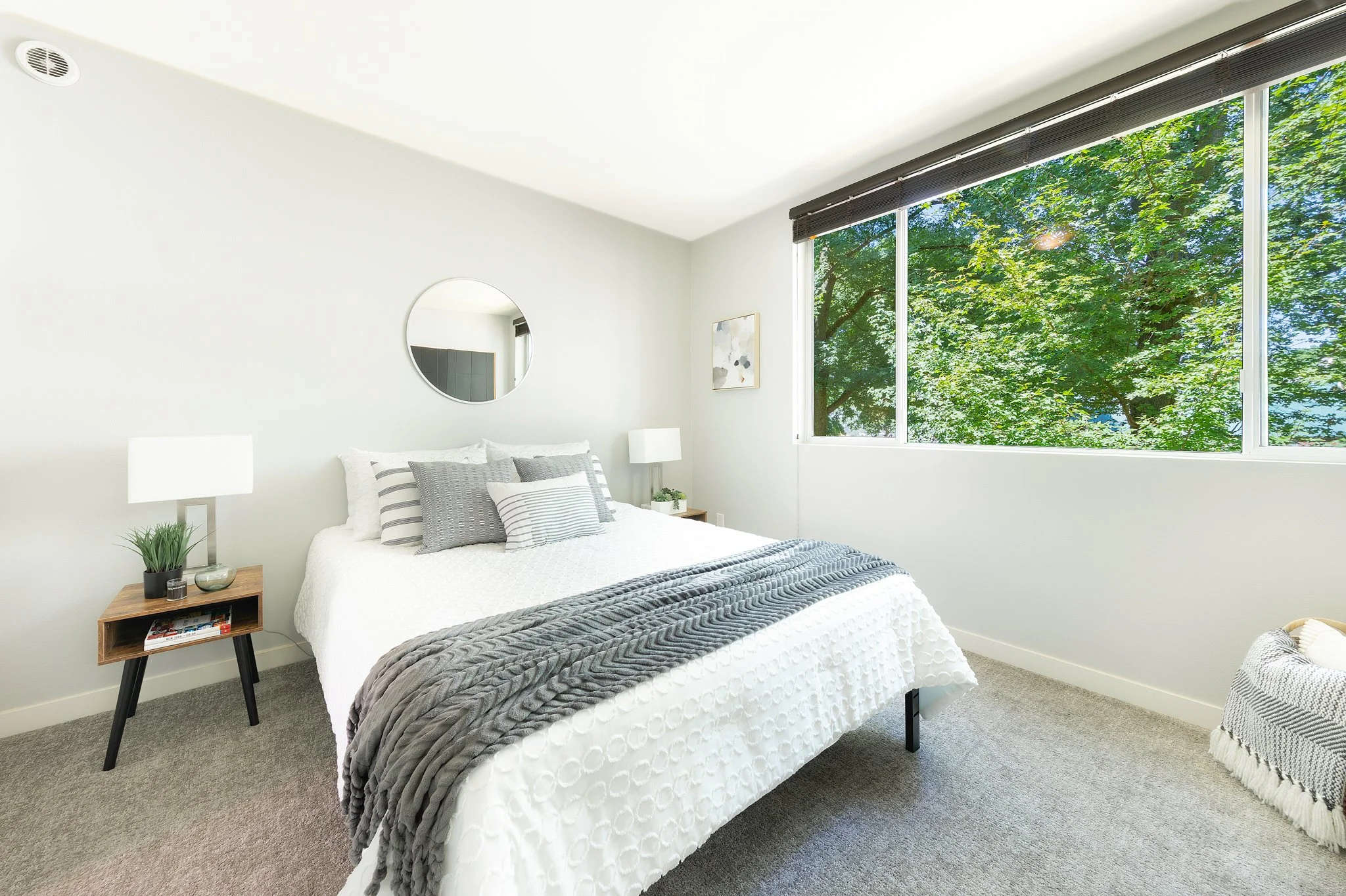 A bedroom with a large window showing green trees outside. The bed has white and gray striped pillows and a gray blanket. There are two white lamps on small wooden nightstands, one on each side of the bed. The room has minimal decor, including a smal