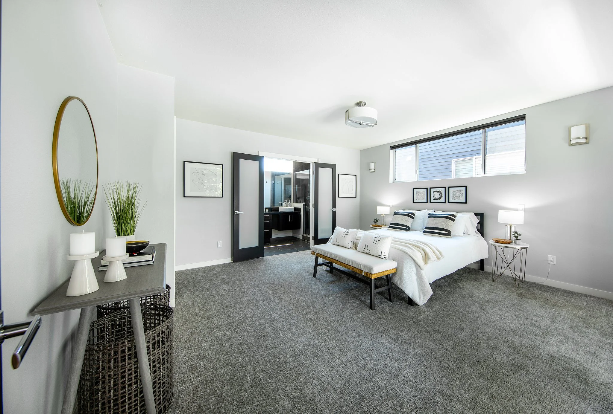 A modern bedroom with a white bed, black headboard, and black and white striped pillows. Art prints hang above the bed, and there are white lamps on nightstands. A small bench sits at the foot of the bed. A window with a view of another building is a