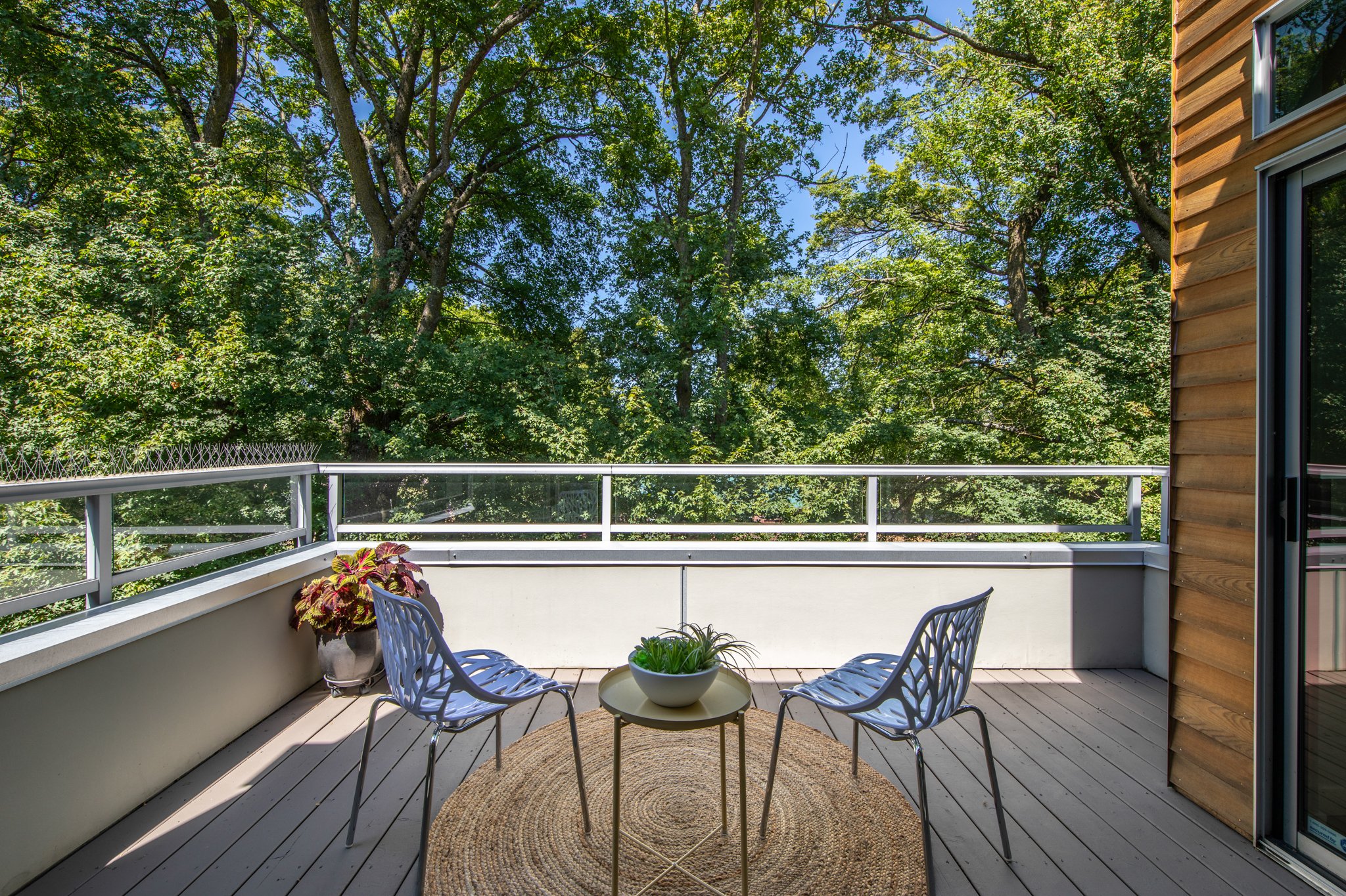 A balcony with a round table and four metal chairs, a potted plant on the table, a rug, and trees in the background with a blue sky.