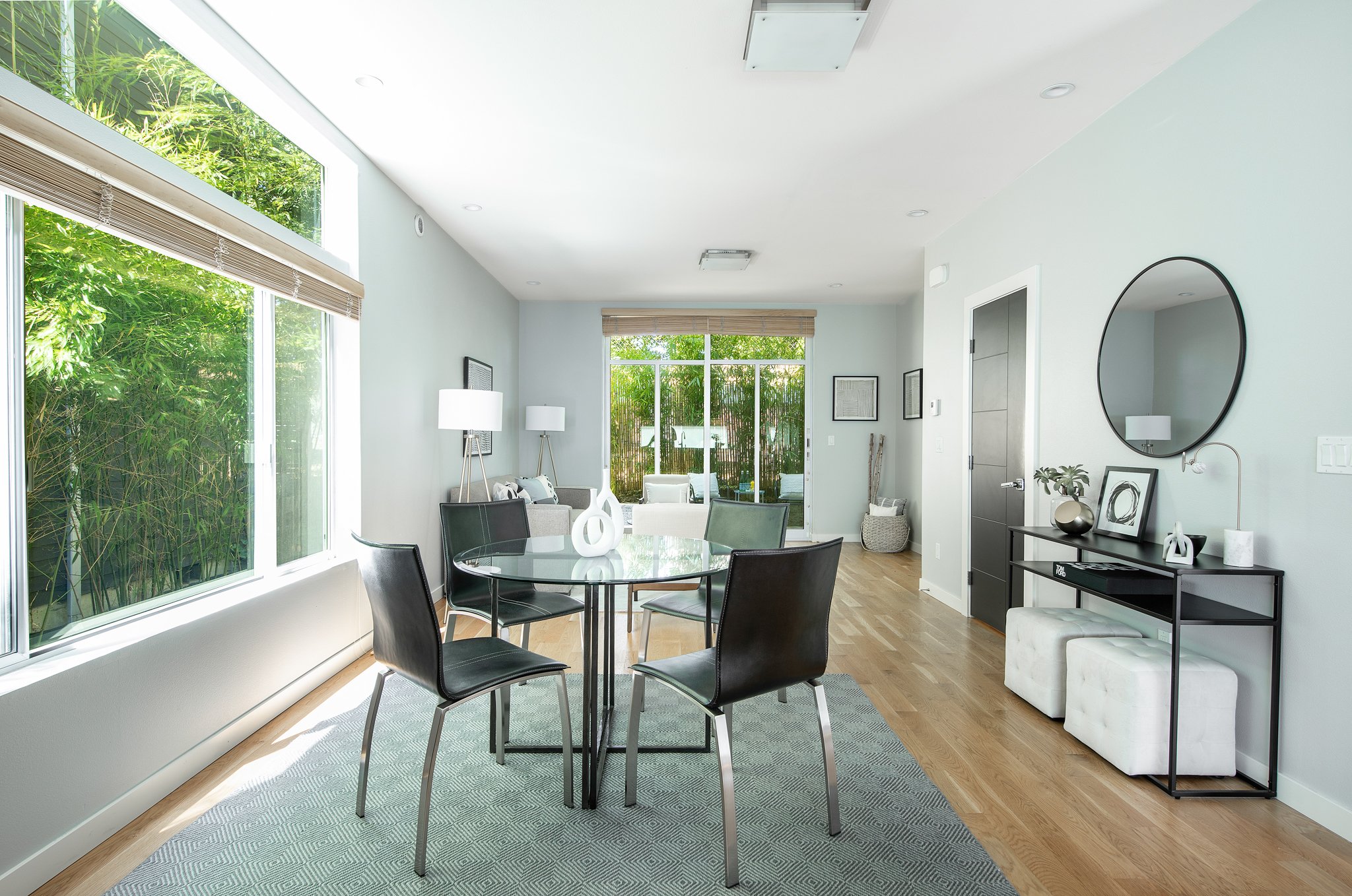 Modern living room with large windows, black dining table with six chairs, white sofas, and decorative items on a black console table, with a large round mirror on the wall.