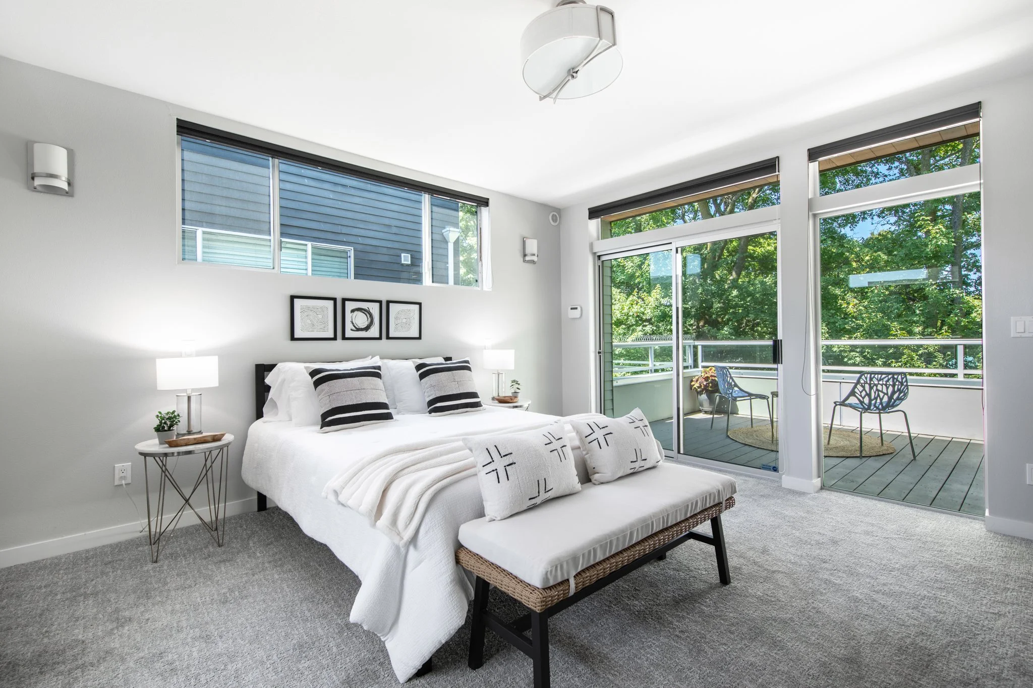 A modern bedroom with a large bed, decorative pillows, nightstands with lamps, wall art, and a balcony with outdoor seating and a view of trees.