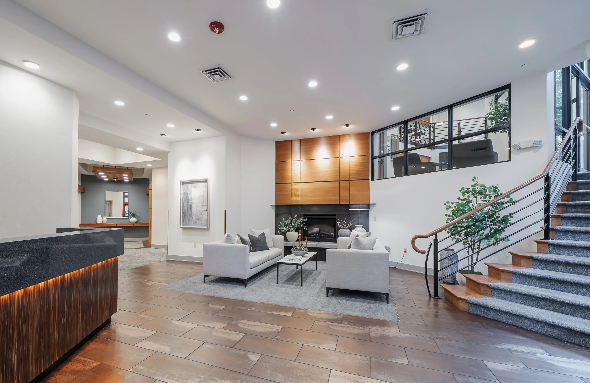 Interior of a modern, well-lit lobby or lounge area with white walls, wooden accents, a fireplace, beige sofas, a glass staircase with metal handrails, and large windows letting in natural light.