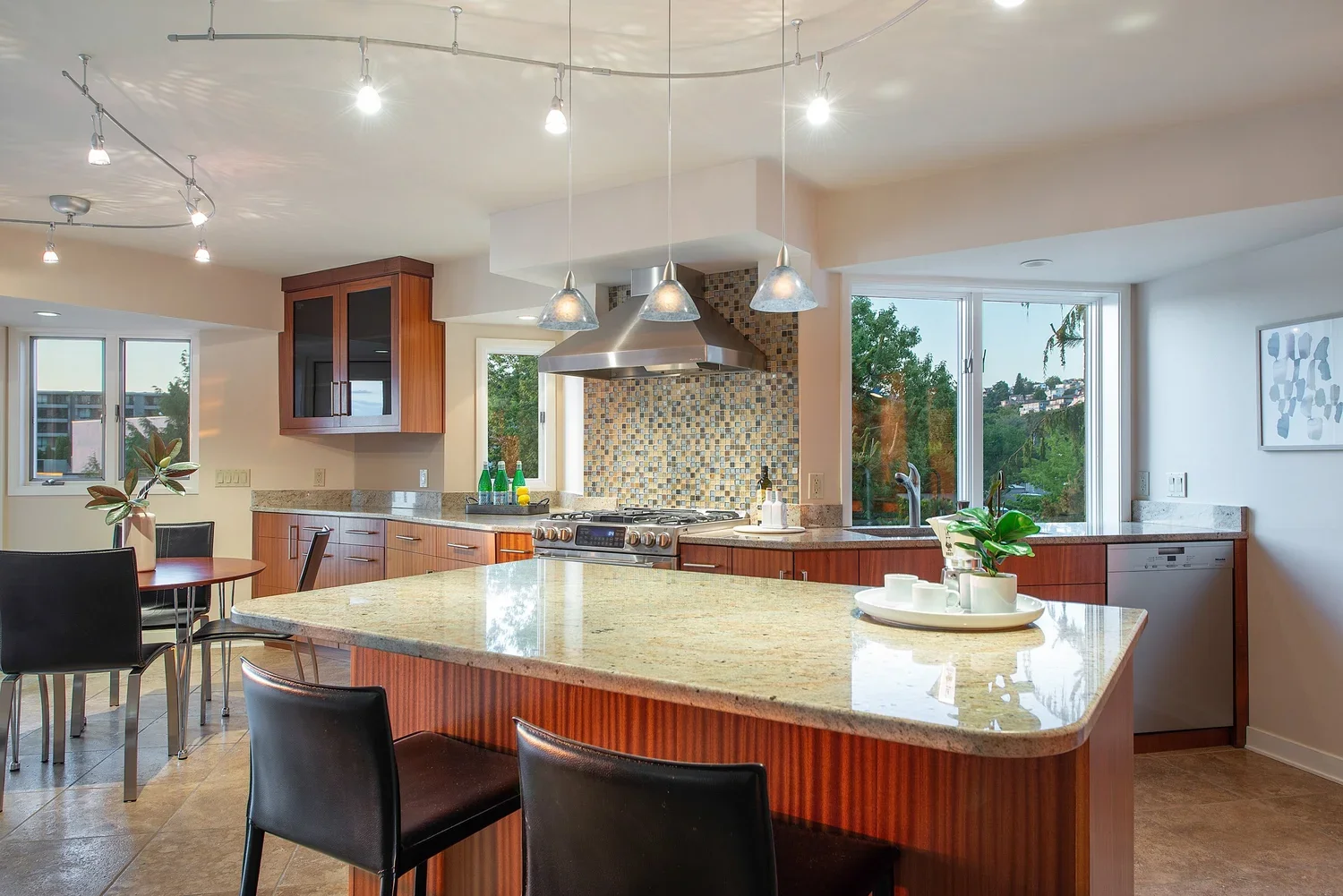 Modern kitchen with granite island, wooden cabinets, colorful mosaic backsplash, stainless steel stove and vent hood, pendant and track lighting, and large windows showing greenery outside.