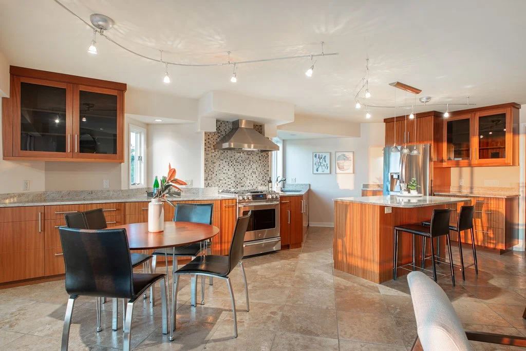 Modern kitchen with wooden cabinets, granite countertops, stainless steel appliances, a dining table with four chairs, and a kitchen island with barstools, illuminated by hanging and ceiling lights, with a tiled backsplash and windows letting in natu