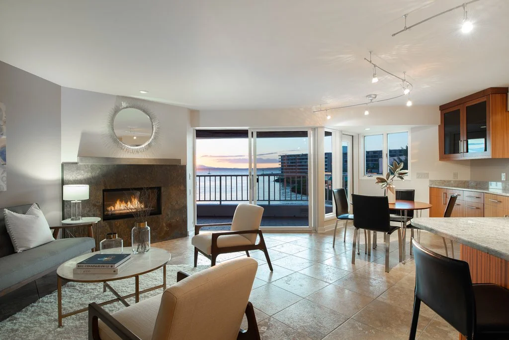Modern living room with a window view of water at sunset, a fireplace, gray sofa, armchairs, dining table with four chairs, and kitchen area with wooden cabinets.