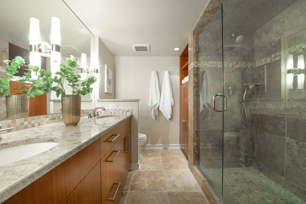A modern bathroom with a dual-sink vanity, large mirror, potted green plant, and a walk-in shower with glass door, gray tiles, and pebble floor. Two white towels hang on hooks in the background.