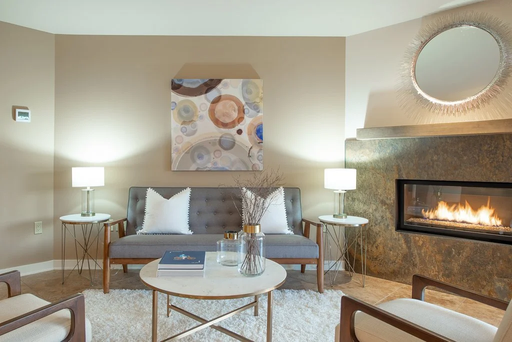 Living room with fireplace, art piece on beige wall, gray sofa with white pillows, round white coffee table with books and vases, two side tables with lamps, round mirror above fireplace.