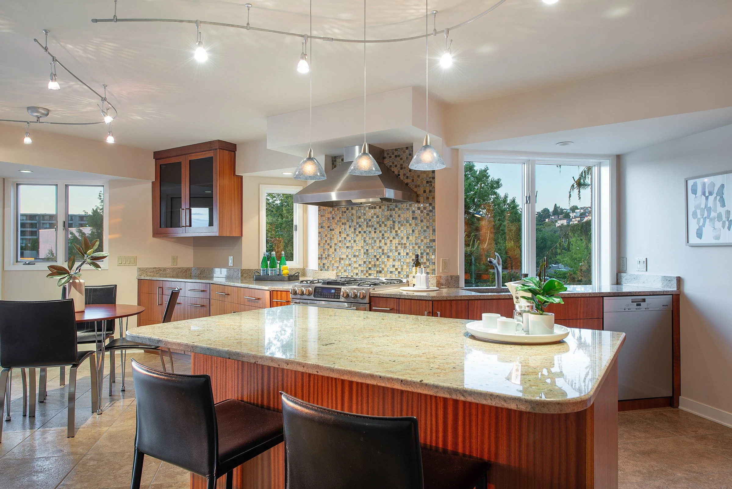 Modern kitchen with wooden cabinets, granite countertops, a stainless steel range hood, colorful tile backsplash, large windows showing trees outside, and a dining table with black chairs.