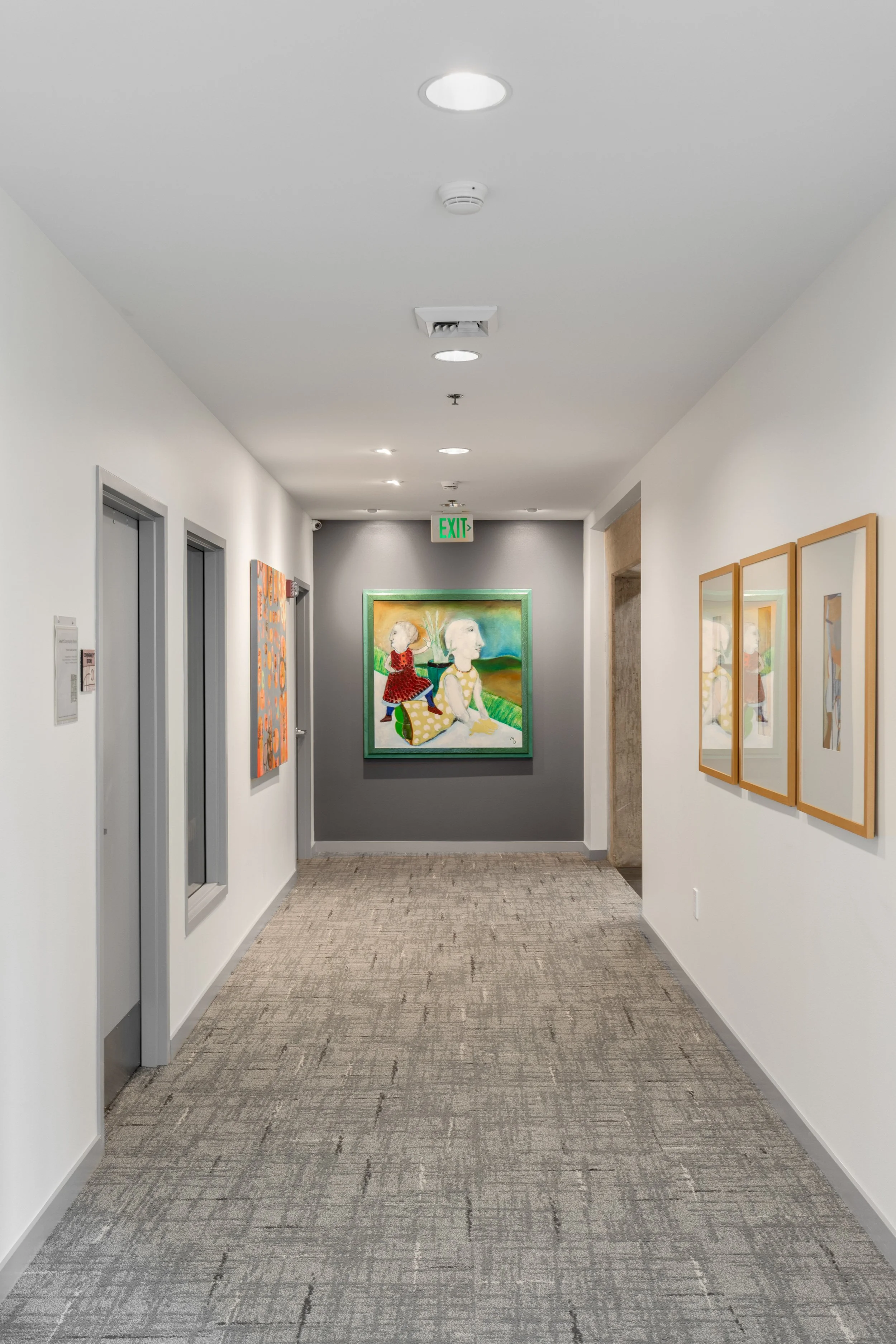 A hallway in a multi-family community with white walls, grey doors, and multiple colorful paintings on display. The hallway has an exit sign on the ceiling, and the floor is carpeted in a neutral tone.