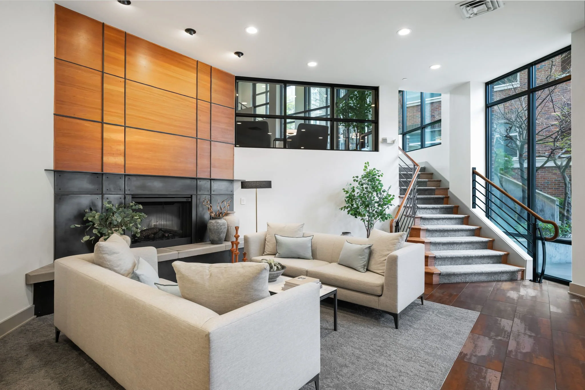 Living room with beige sofas, a fireplace, a coffee table, large windows, a staircase, and indoor plants with trees visible outside.