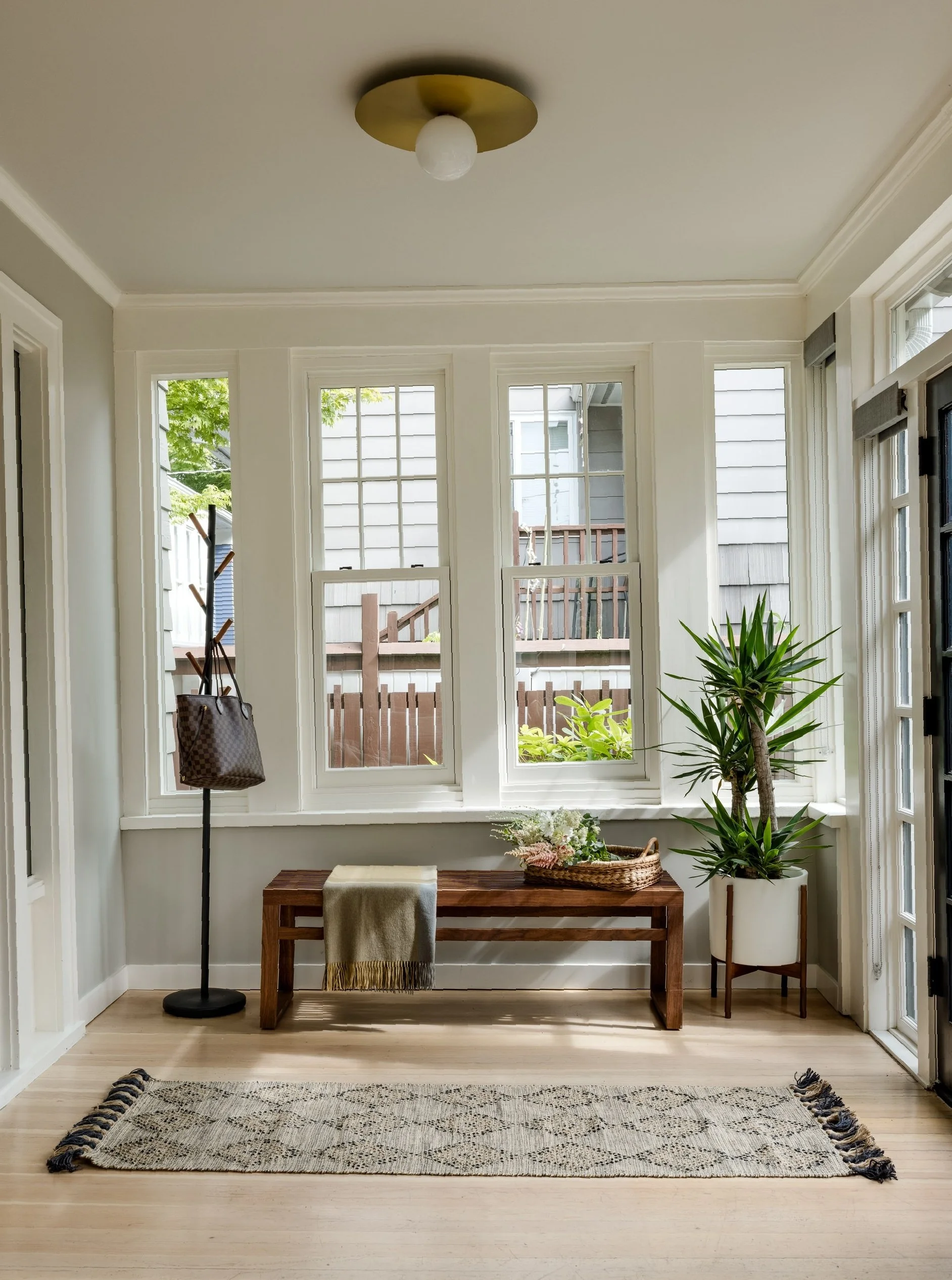 Sunroom with large windows, a wooden bench with a blanket, a potted plant, a rug, a coat rack, and a ceiling light.