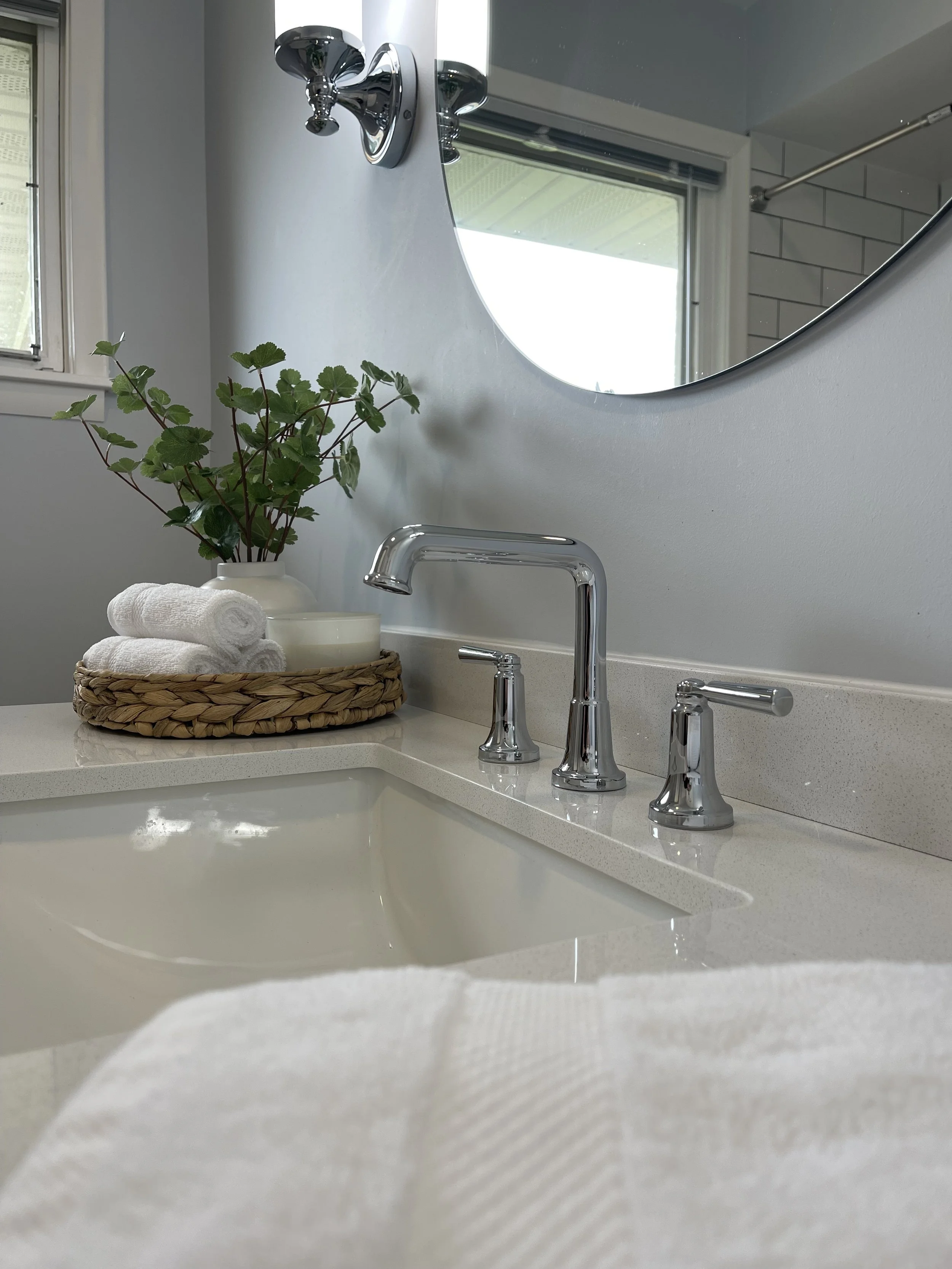 Bathroom sink with two silver faucets, a mirror above, a window on the left, and a basket with rolled white towels and a leafy green plant on the counter.
